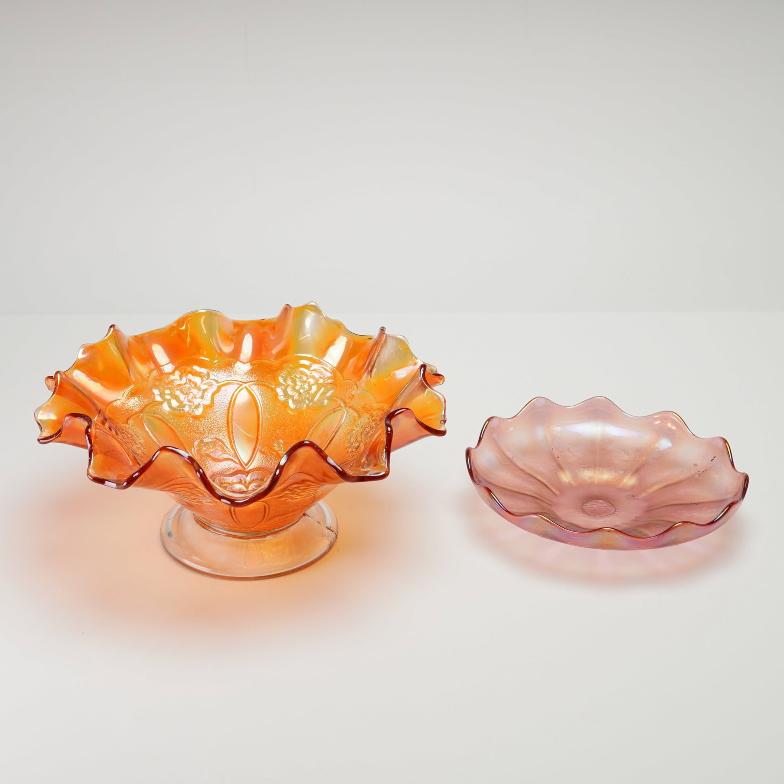 Two Piece Glass Bowl and Dish Group Including Dugan Marigold Carnival Glass and Pink Hand Blown: This two piece glass bowl and dish group features iridescent finishes. The larger item is an orange Dugan Double Stem Rose carnival glass bowl with a ruffled edge and footed base. Its interior is deco