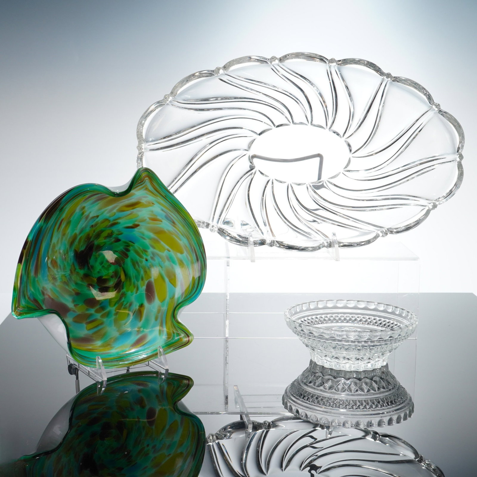 Three Pieces of Decorative Glassware Bowls and Tray: This lot consists of three pieces of decorative glassware. The first item is a clear glass oval platter featuring a radiating spiral optic pattern extending from a central well to a scalloped rim, cha