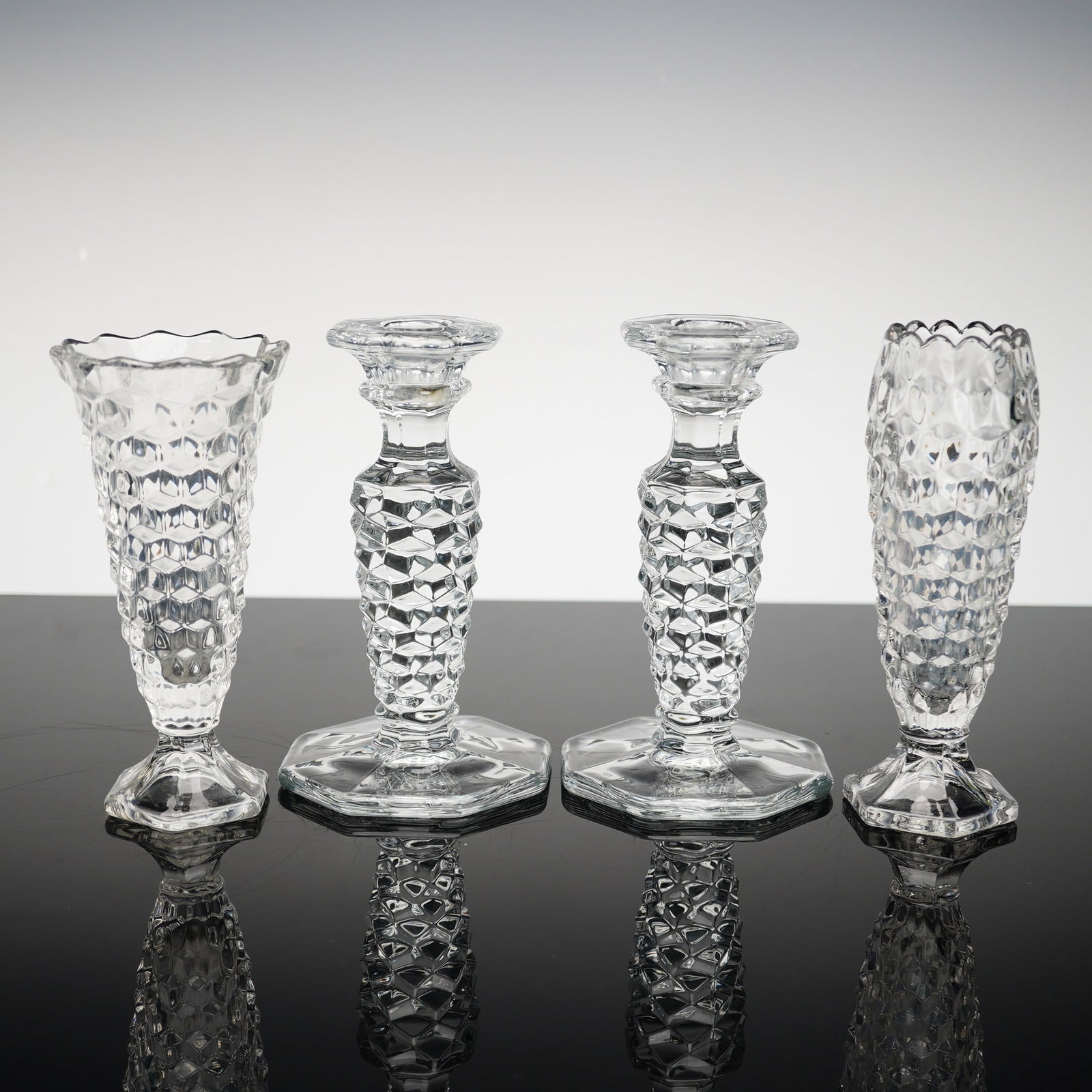Fostoria American Pattern Clear Glass Candlesticks and Bud Vases: Produced by the Fostoria Glass Company, this four-piece set is crafted from clear press-molded crystal in the celebrated American pattern, also known as Line 2056. This mid-20th-century design is famo