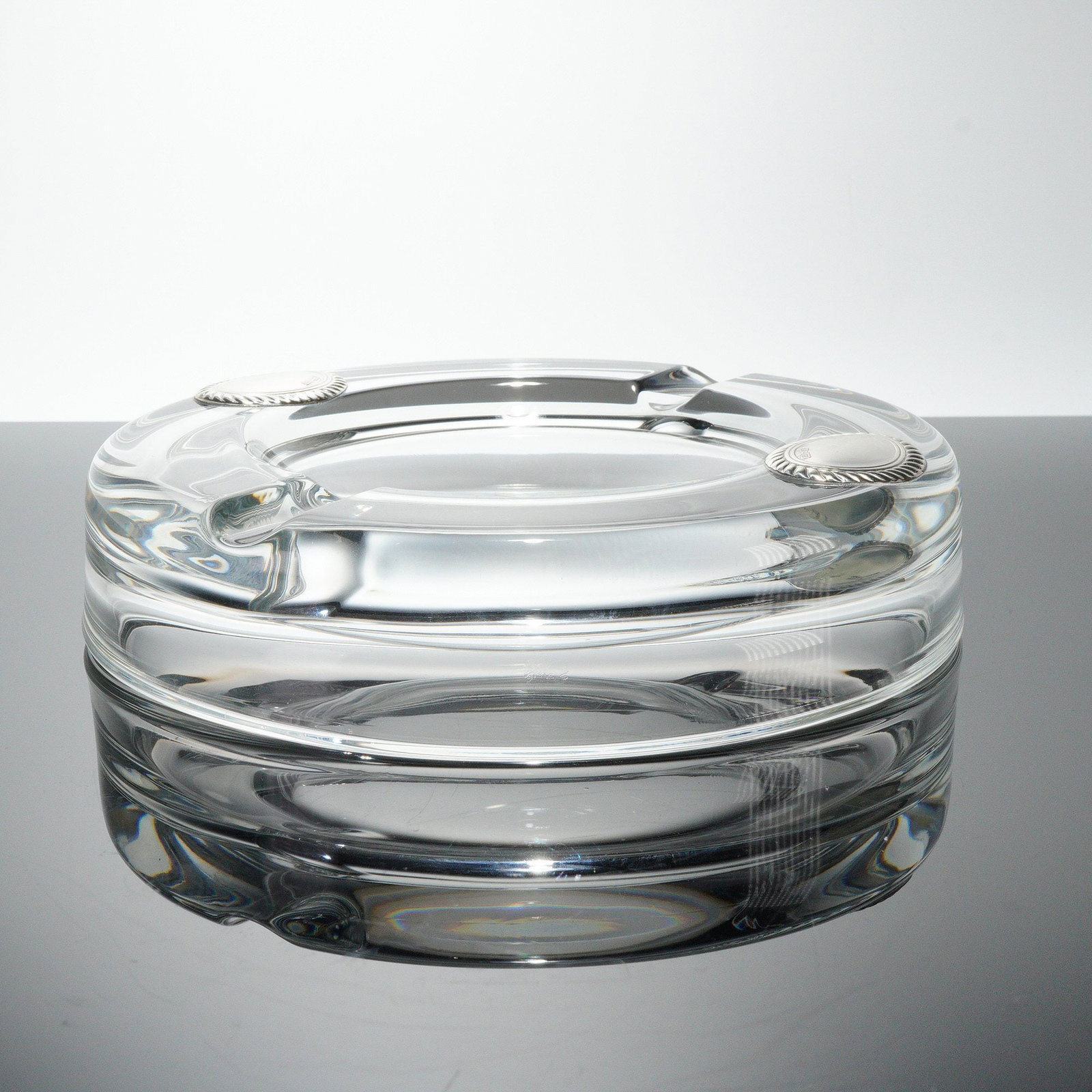 Glass Ashtray with Silver Toned Medallions: A circular ashtray crafted from heavy clear glass. The contemporary design features a thick body with a recessed central bowl and four molded cigarette rests around the rim. The object is accented by 