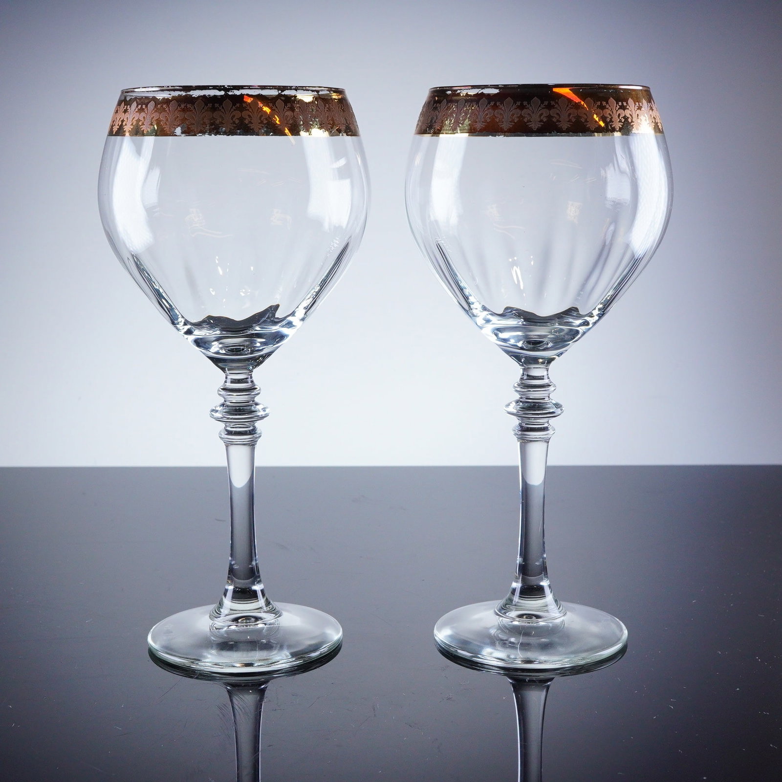 2pc Gold Rim Fleur De Lis Wine Glasses: A pair of stemmed clear crystal wine glasses featuring a classical revival style with tall, knopped stems and slightly tapered, tulip-shaped bowls. The bowls are detailed with vertical facet cutting e