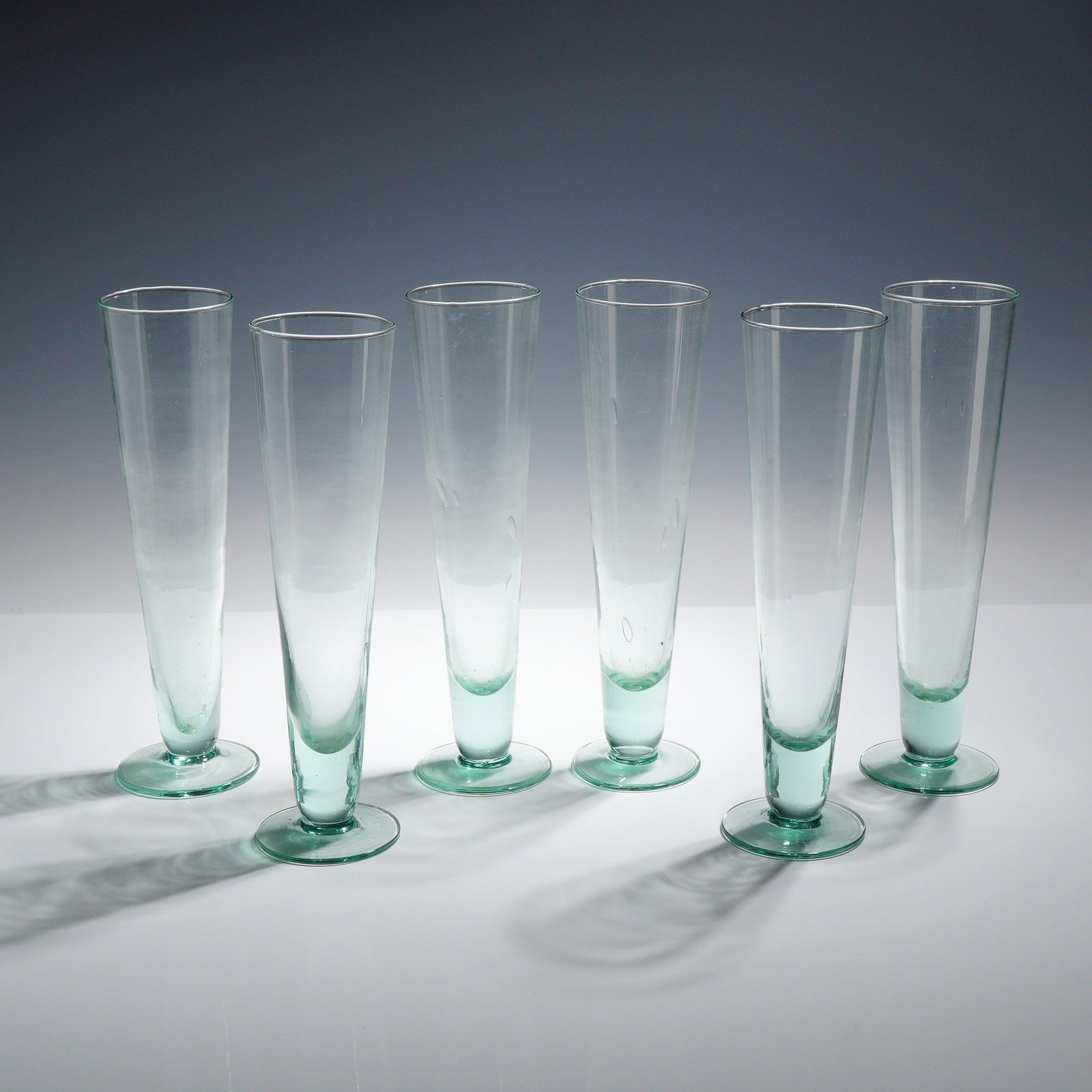 6pc Hand-Blown Pale Green Pilsner Glasses: A set of 6 hand-blown pilsner-style glasses or tall tumblers. Each vessel is constructed with a tall, slightly tapered cylindrical body joined to an applied circular disc foot. The glass is characteri