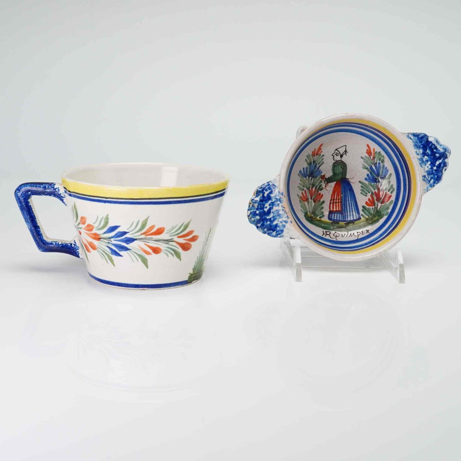 Two French Henriot Quimper Hand Painted Faience Pottery Items: This lot consists of two pieces of hand-painted French faience pottery from Quimper. The first item is a tapered ceramic cup with a square-profile handle. The exterior decoration features a floral spr