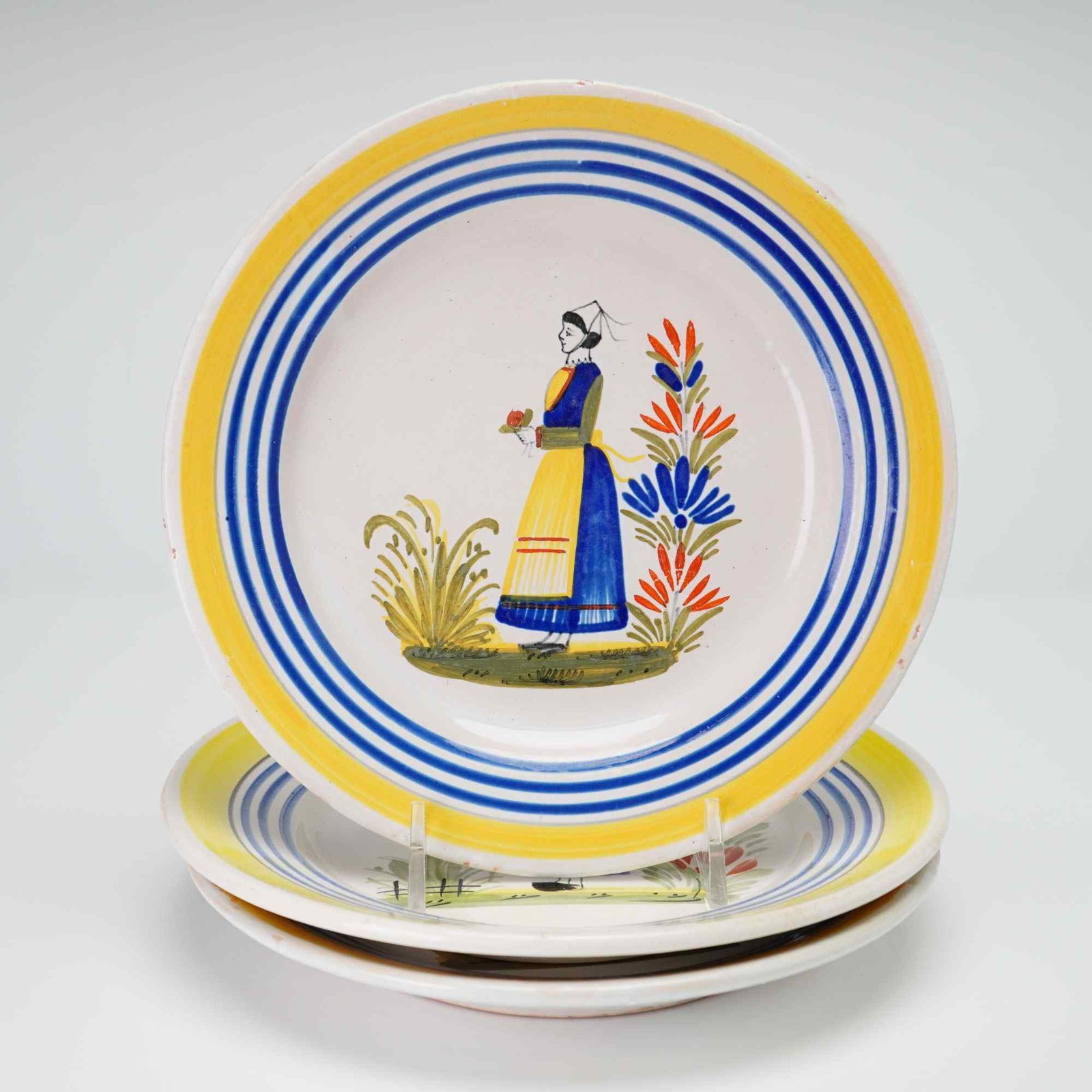 Three Henriot Quimper Hand Painted Faience Breton Woman Plates: This trio of circular plates is crafted from tin-glazed earthenware, commonly known as faience, produced by Henriot Quimper in Brittany, France. Each plate features a hand-painted central motif of a B