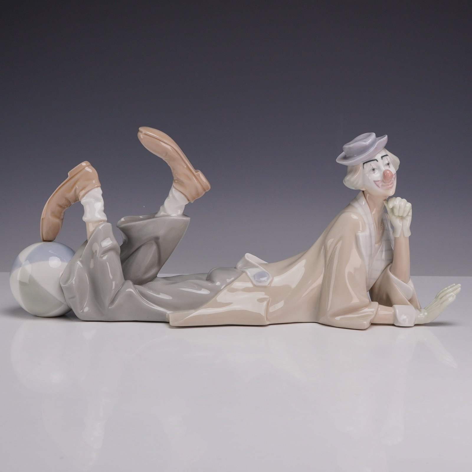 Lladro Porcelain Figurine Clown 01004618: The piece depicts a clown in a pensive reclining pose, resting on his stomach with his chin propped in his right hand. His legs are kicked upward behind him, with his left foot balanced on a large, pa