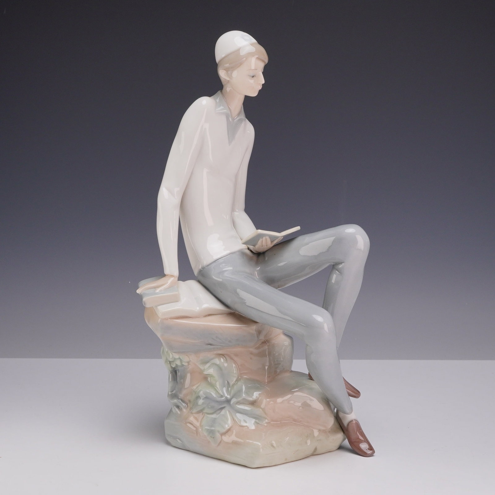 Lladro Glazed Porcelain Figurine, Hebrew Student 01004684: This piece, model number 4684, was designed by sculptor Juan Huerta. The figure depicts a young man wearing a white kippah, a cream-colored long-sleeve shirt, and grey trousers. He is seated on a styl