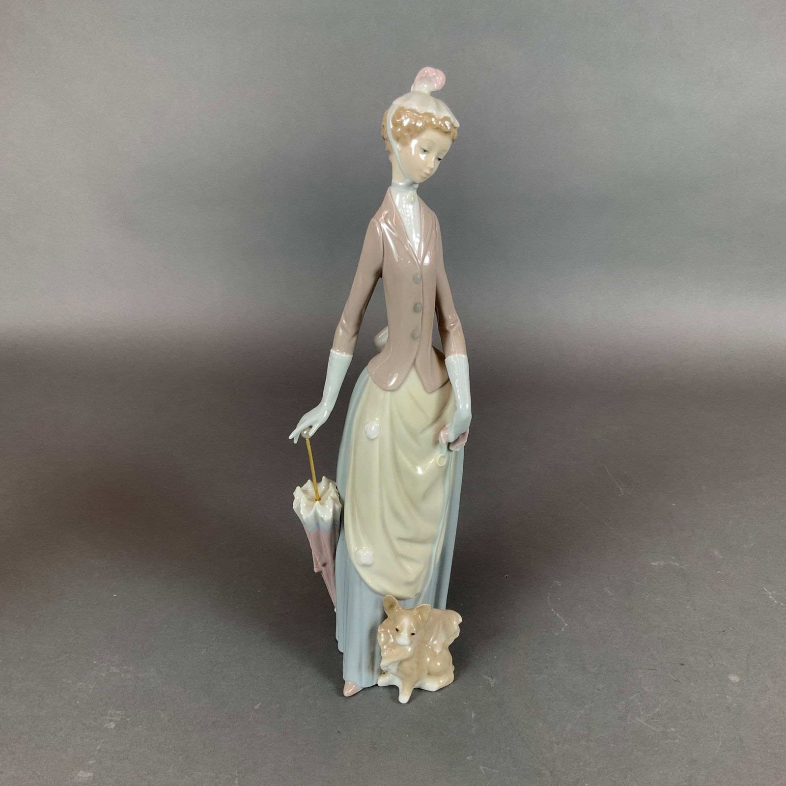 Lladro Glazed Porcelain Figurine Woman with Dog Model 4761: This is a glazed porcelain figurine depicting a woman in Edwardian-style attire, wearing a mauve jacket with grey buttons over a pale blue pleated skirt with a cream-colored apron front. She is accent