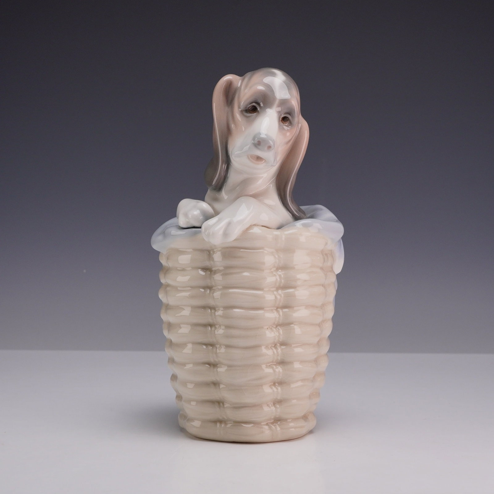 Lladro Porcelain Figurine Dog in a Basket 01001128: This Lladro glazed porcelain figurine depicts a basset hound puppy with long ears peering out from a woven-texture laundry basket. The puppy is decorated with grey and tan patches, while a pale blue c