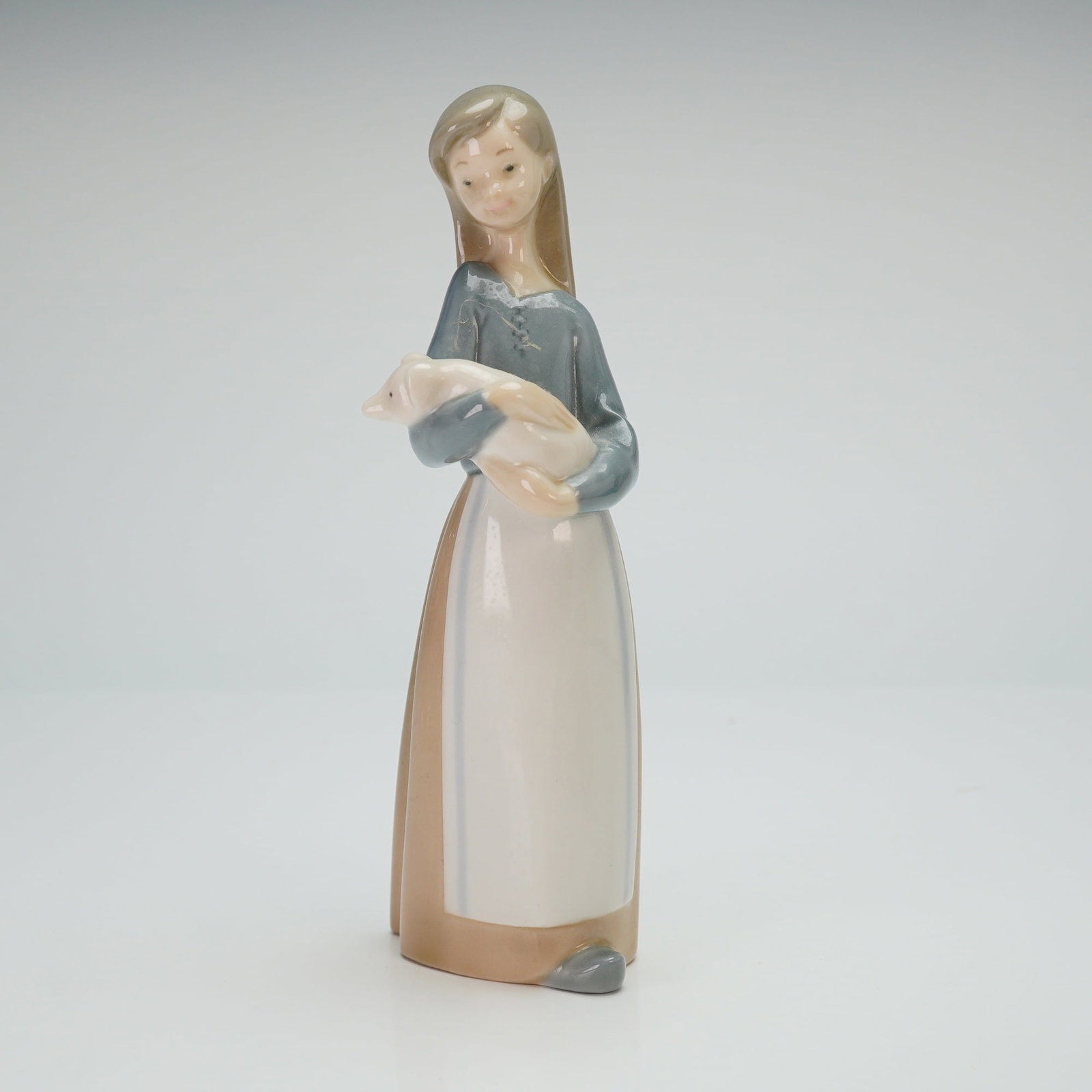Lladro Porcelain Figurine Girl with Pig 01001011: The piece depicts a young woman standing and cradling a small piglet. She is rendered in a muted color palette, wearing a long tan dress, a blue-gray bodice, and a white apron. The figurine features a