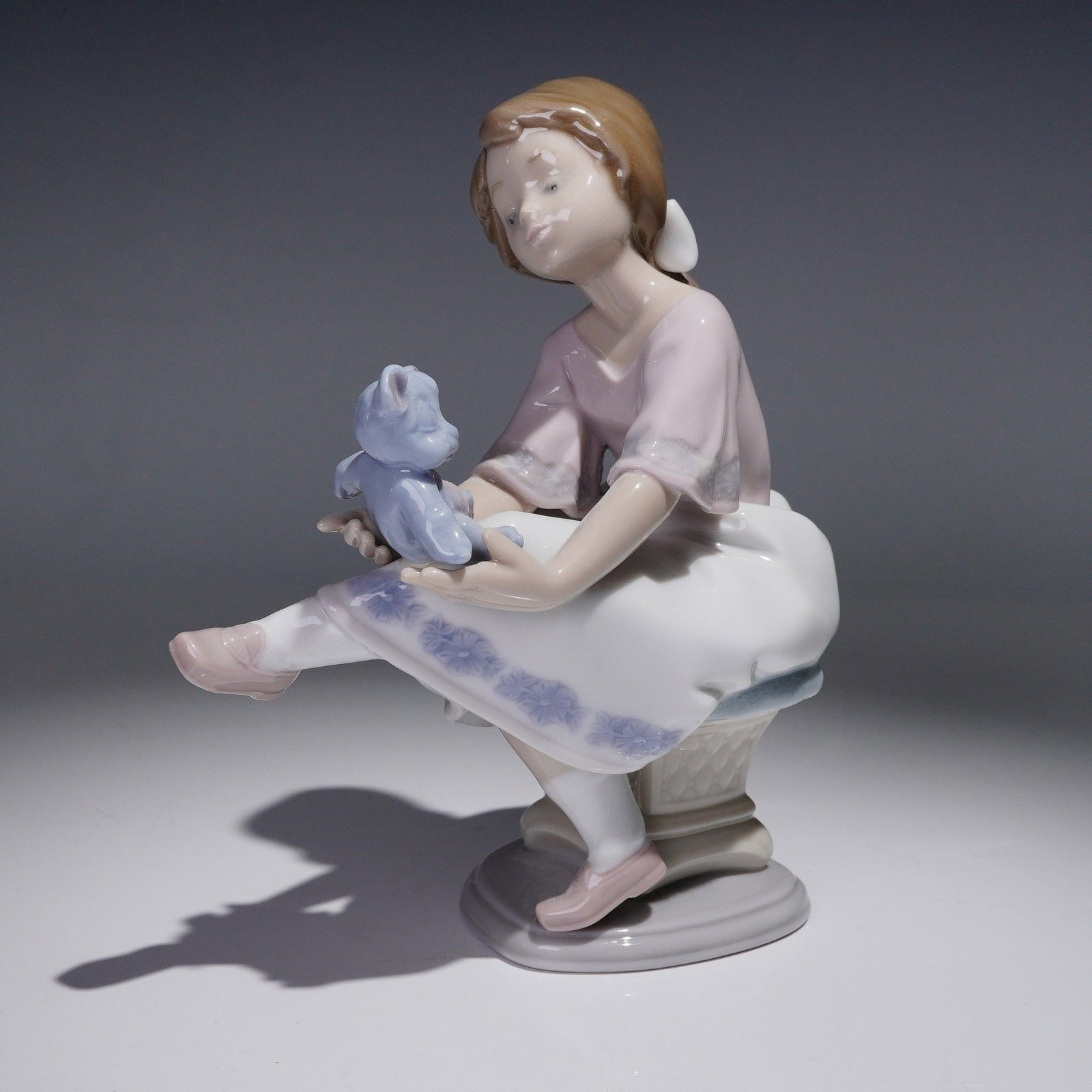 Lladro Porcelain Figurine, Best Friends 1007620: Depicting a young girl seated on a pedestal-style base while holding a plush teddy bear in her outstretched hands. The girl wears a light pink blouse, white skirt with molded lavender floral detail, a