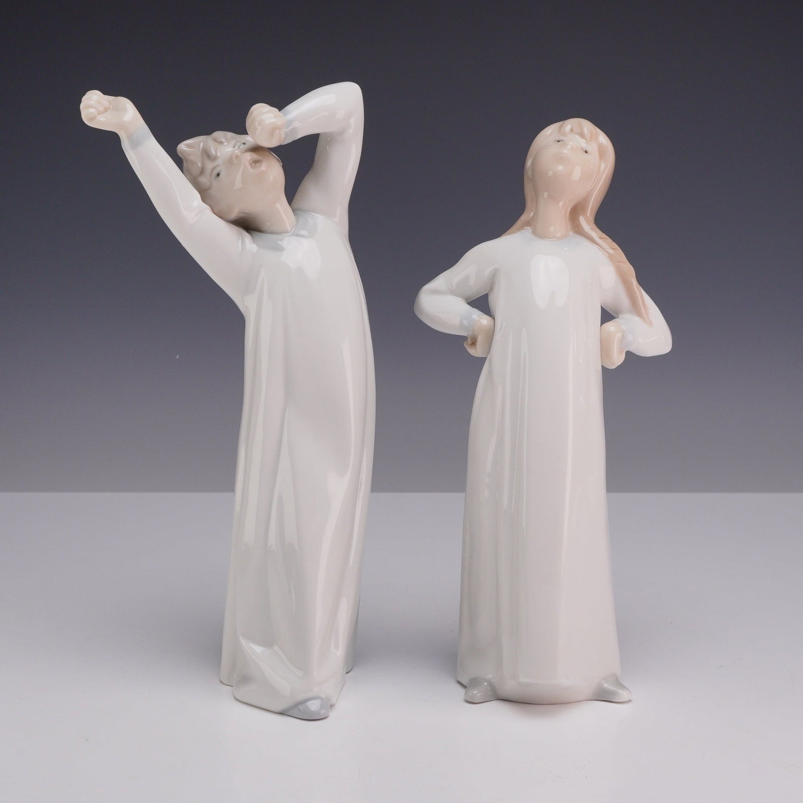 Two Lladro Porcelain Figurines, Boy Yawning 01004870 and Girl Stretching 01004872: Both figures are rendered in glazed porcelain with a soft pastel color scheme consisting of white, gray, and beige tones. The Boy Awakening figure, model number 4870, depicts a young boy in a long whi