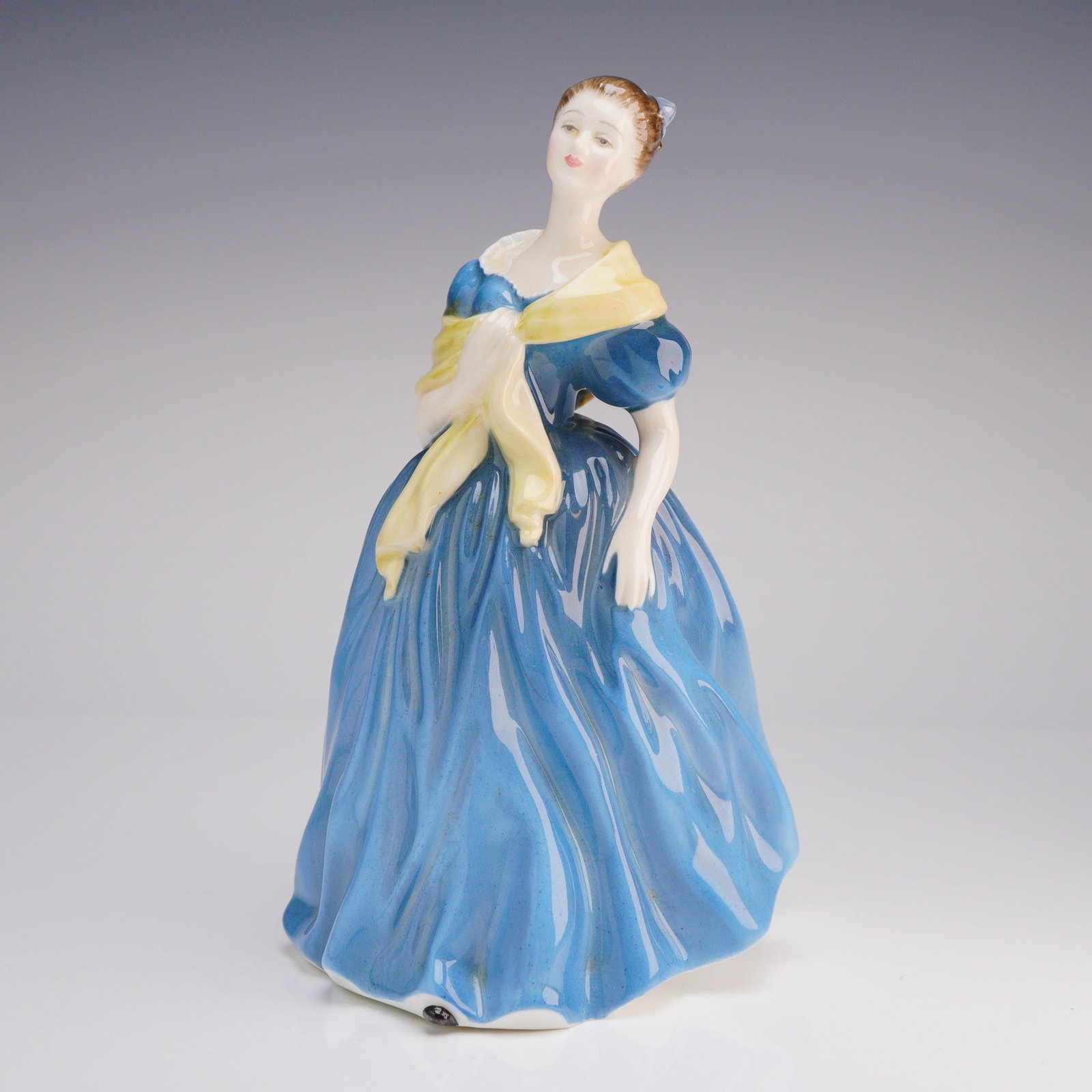 Royal Doulton Adrienne Figurine HN 2304: The figurine depicts a standing woman dressed in a flowing, voluminous blue gown and a pale-yellow shawl draped around her shoulders, stylized in a mid-20th-century decorative manner. The base feature