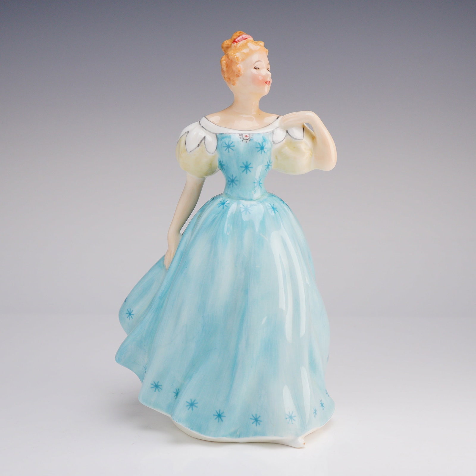 Royal Doulton Figurine Enchantment HN2178: The figure depicts a woman with reddish hair styled in an updo, wearing a voluminous, flowing light blue ballgown. The dress features white puff sleeves and a white ruffled hem, decorated throughout w