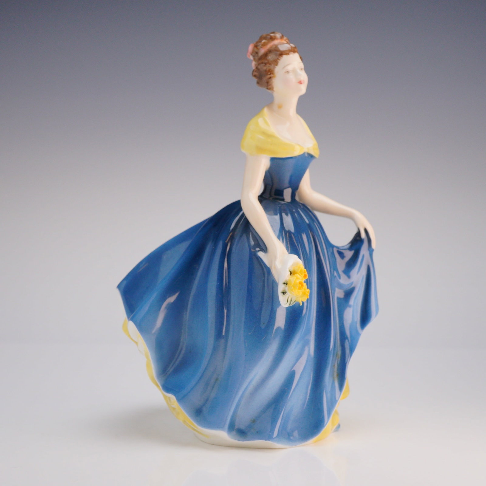 Royal Doulton Figurine Melanie HN2271: Figurine depicts a woman in a full blue gown accented with a pale-yellow off-the-shoulder shawl and yellow ruffles visible beneath the skirt. The figure holds a small bouquet of yellow roses. The piec