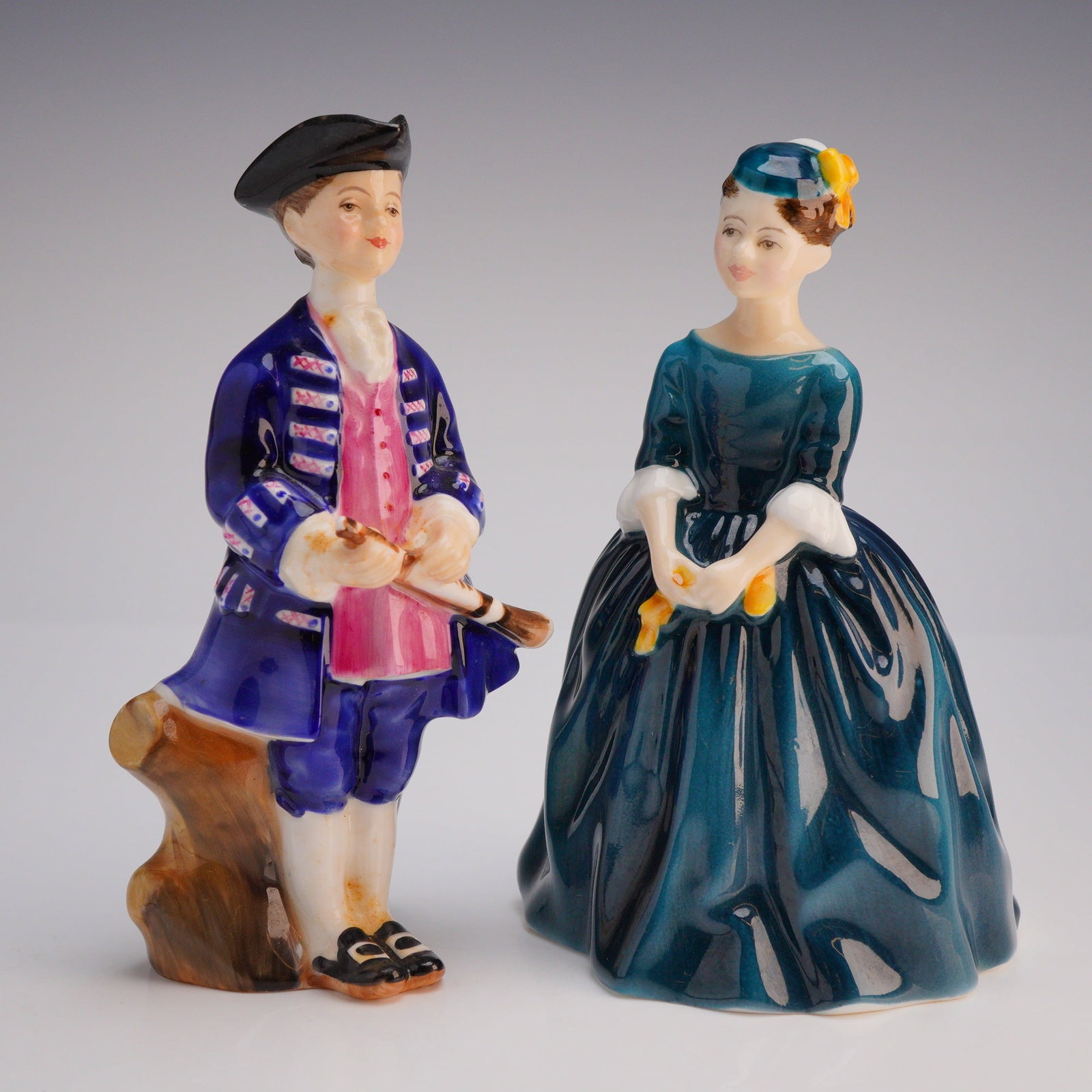 2pc Royal Doulton Figurines Cherie HN2341 and Boy from Williamsburg HN2183: The pieces depict a historical costumed couple rendered in a high-gloss polychrome glaze featuring deep cobalt blue, teal, and pink accents. The male figure, Boy from Williamsburg, wears a tricorn hat