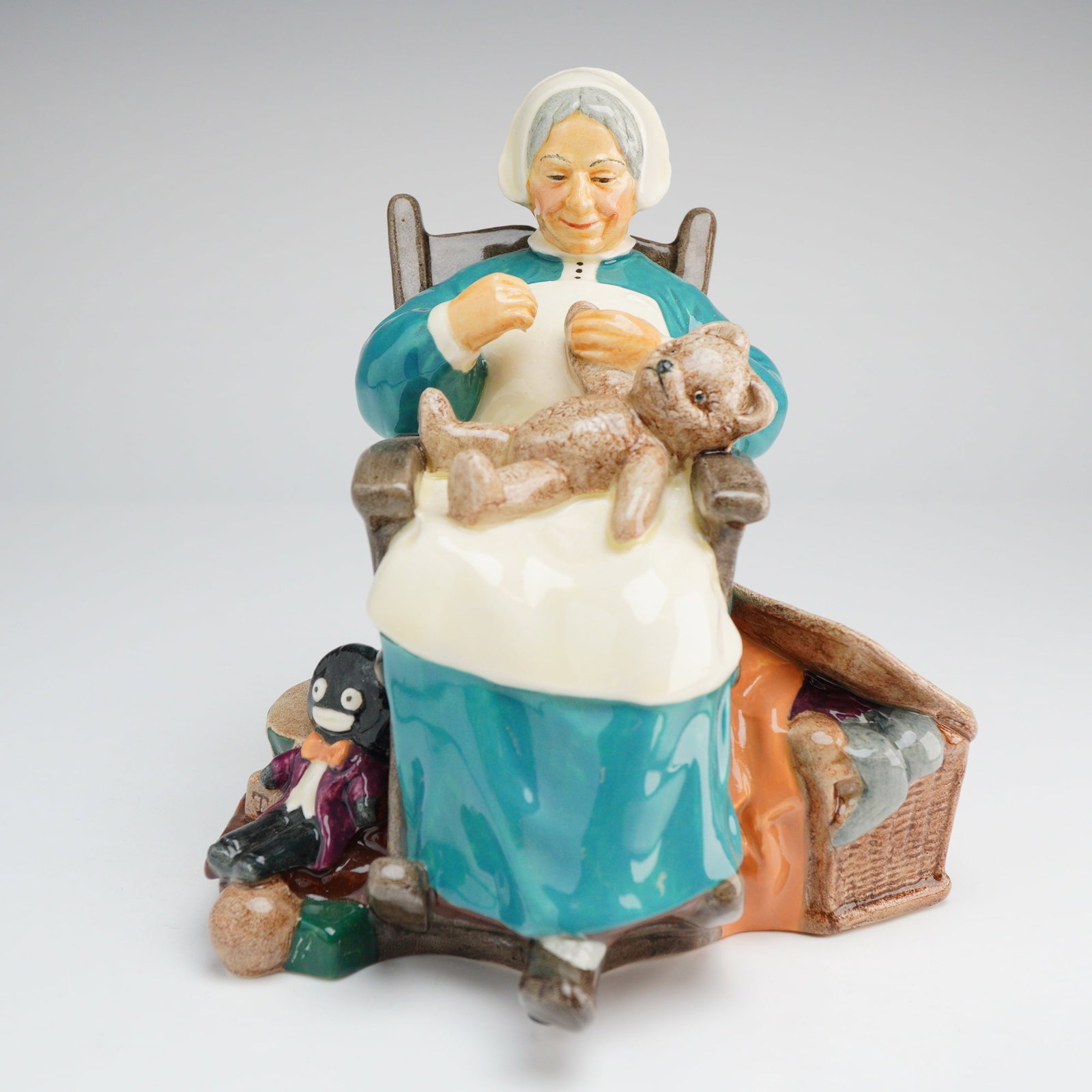 Royal Doulton Figurine, Nanny HN 2221: This Royal Doulton porcelain figurine, titled Nanny (H.N. 2221), depicts an elderly woman seated in a brown rocking chair. The figure is dressed in a teal gown with a white apron and cap, shown in the