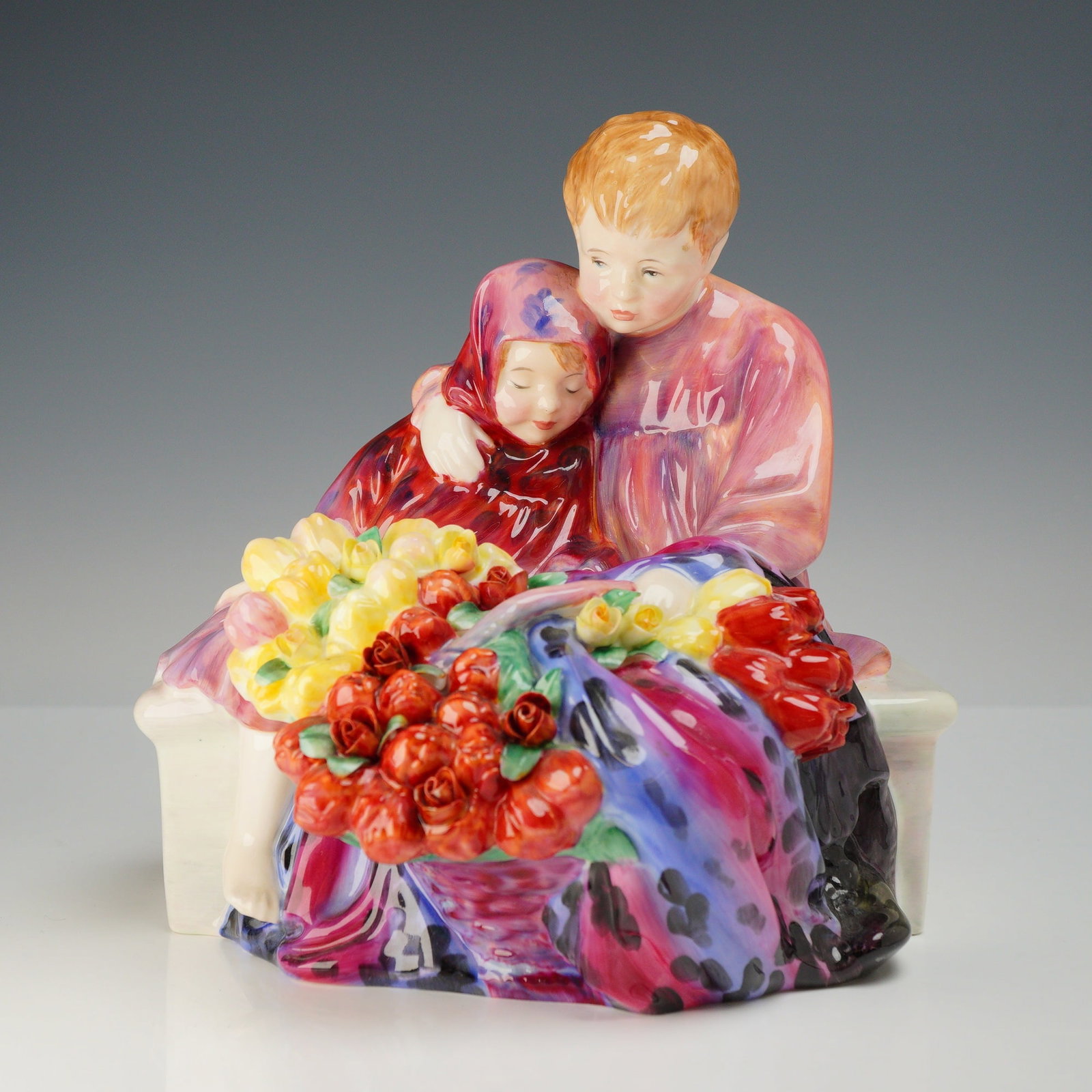 Royal Doulton Bone China Figurine, Flower Sellers Children HN1342: Depicts a young boy and girl seated behind baskets of red roses and yellow tulips. The boy has his arm around the girl's shoulder. The underside of the base features the Royal Doulton backstamp. Dimen