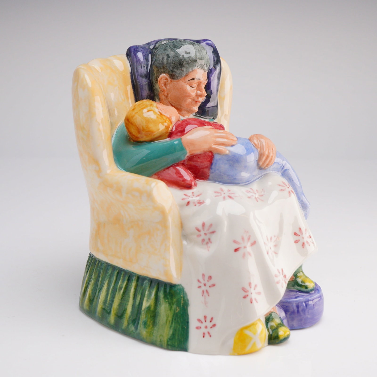 Royal Doulton Figurine Sweet Dreams HN2380: The piece depicts an older woman seated in a high-backed, butter-yellow armchair, cradling a sleeping child in her lap. The figurine is rendered in vibrant polychrome enamels, featuring a white skirt 