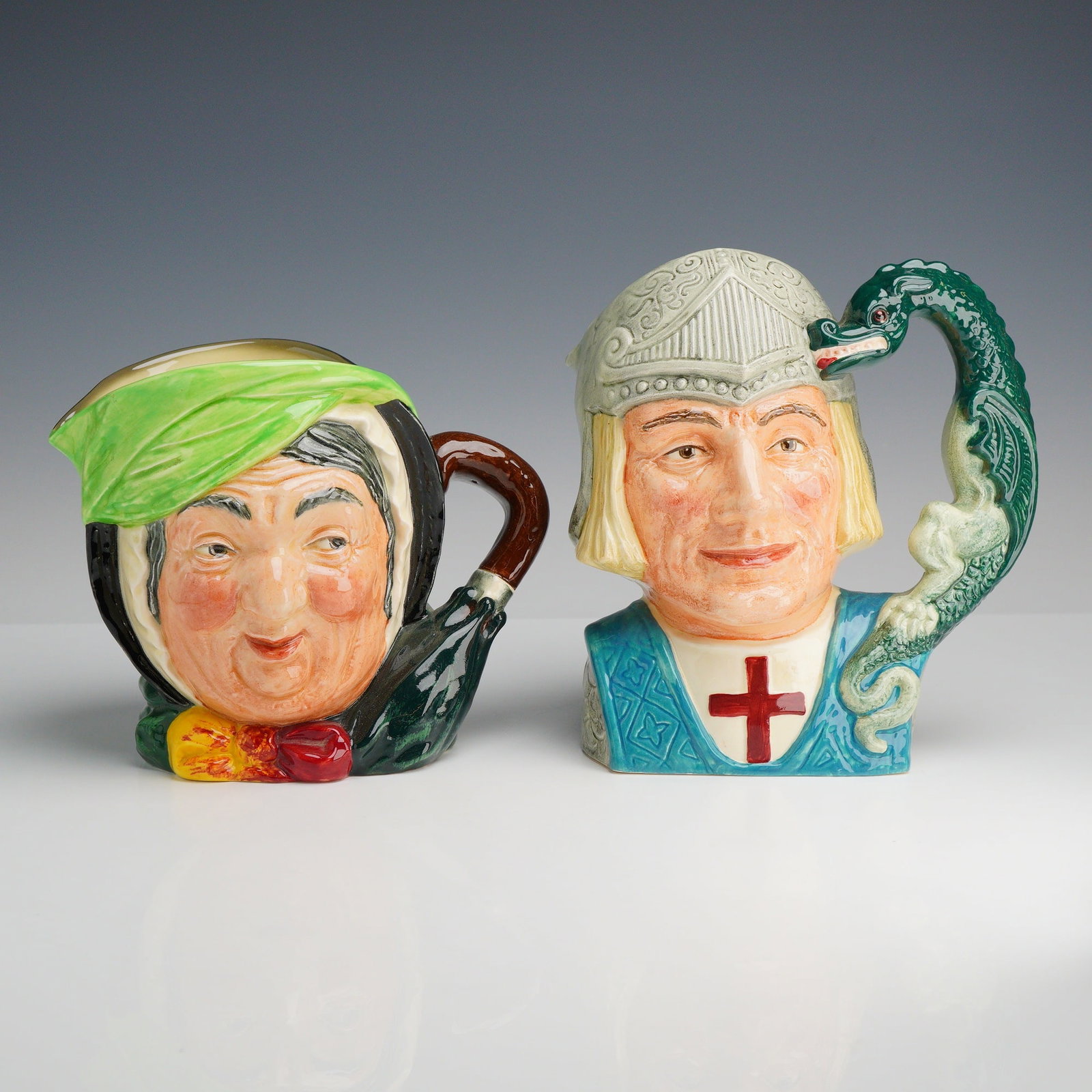 2pc Royal Doulton Character Jugs, Sairey Gamp and St George D6618: The first jug depicts Sairey Gamp, the well-known character from the Charles Dickens novel Martin Chuzzlewit. She is portrayed wearing a black cloak and a bright green bonnet secured with a yellow and