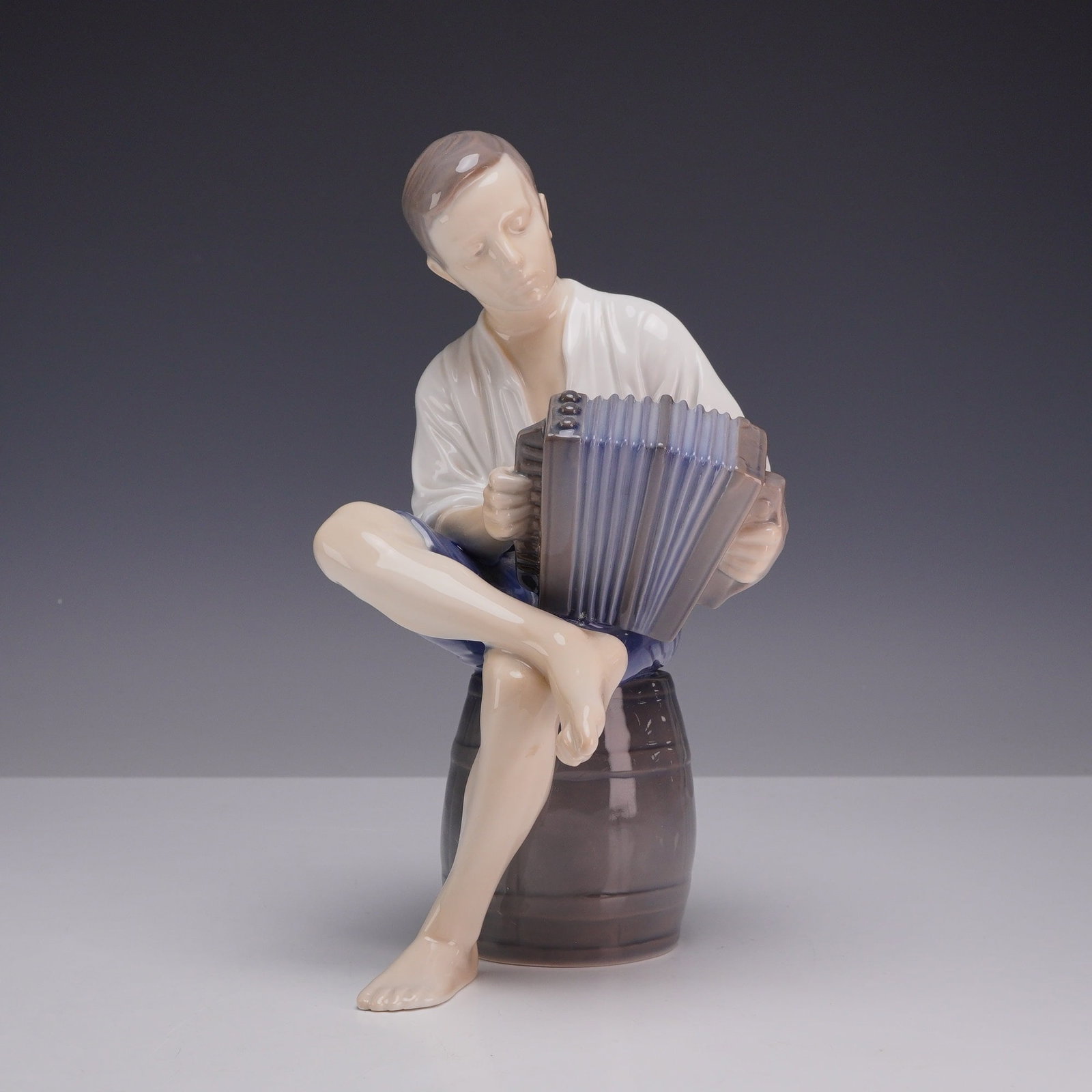 Bing and Grondahl Denmark Porcelain Figurine, Accordion Player: The figure depicts a young man dressed in a white shirt and blue shorts, seated barefoot on a wooden barrel while playing a concertina. The piece is decorated with underglaze hand-painted colors in mu