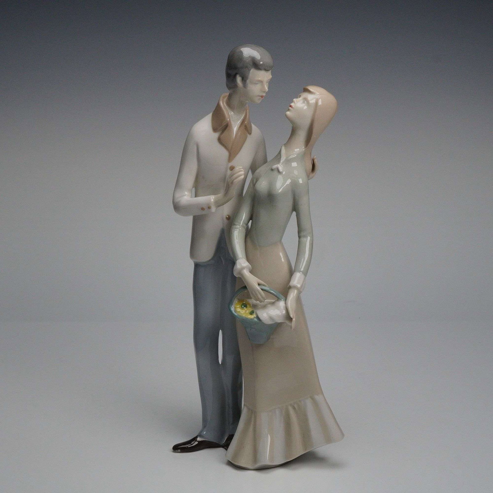 GDR Lippelsdorf Young Couple Porcelain Figurine: A glazed porcelain figurine from Lippelsdorf in the German Democratic Republic depicting a young couple standing closely together, the woman holding a small basket of yellow flowers and dressed in a l