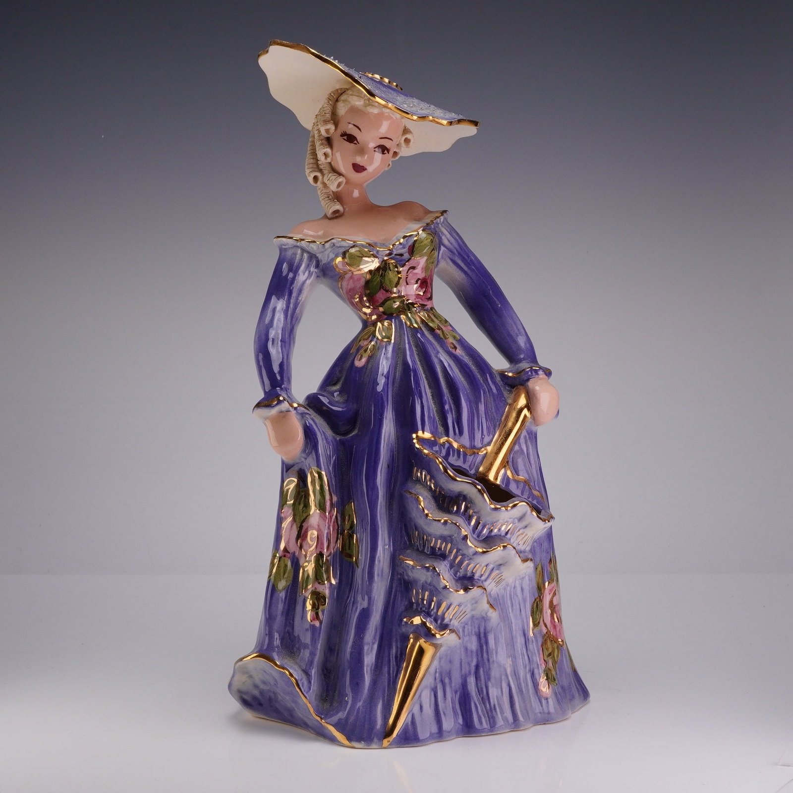 Hedi Schoop California Pottery Glazed Figurine Vase, Woman in Purple Gown: The figure depicts a woman in mid-19th-century style dress, wearing a purple gown decorated with hand-painted pink floral motifs and gold-toned gilded accents. She is modeled with a wide-brimmed hat a