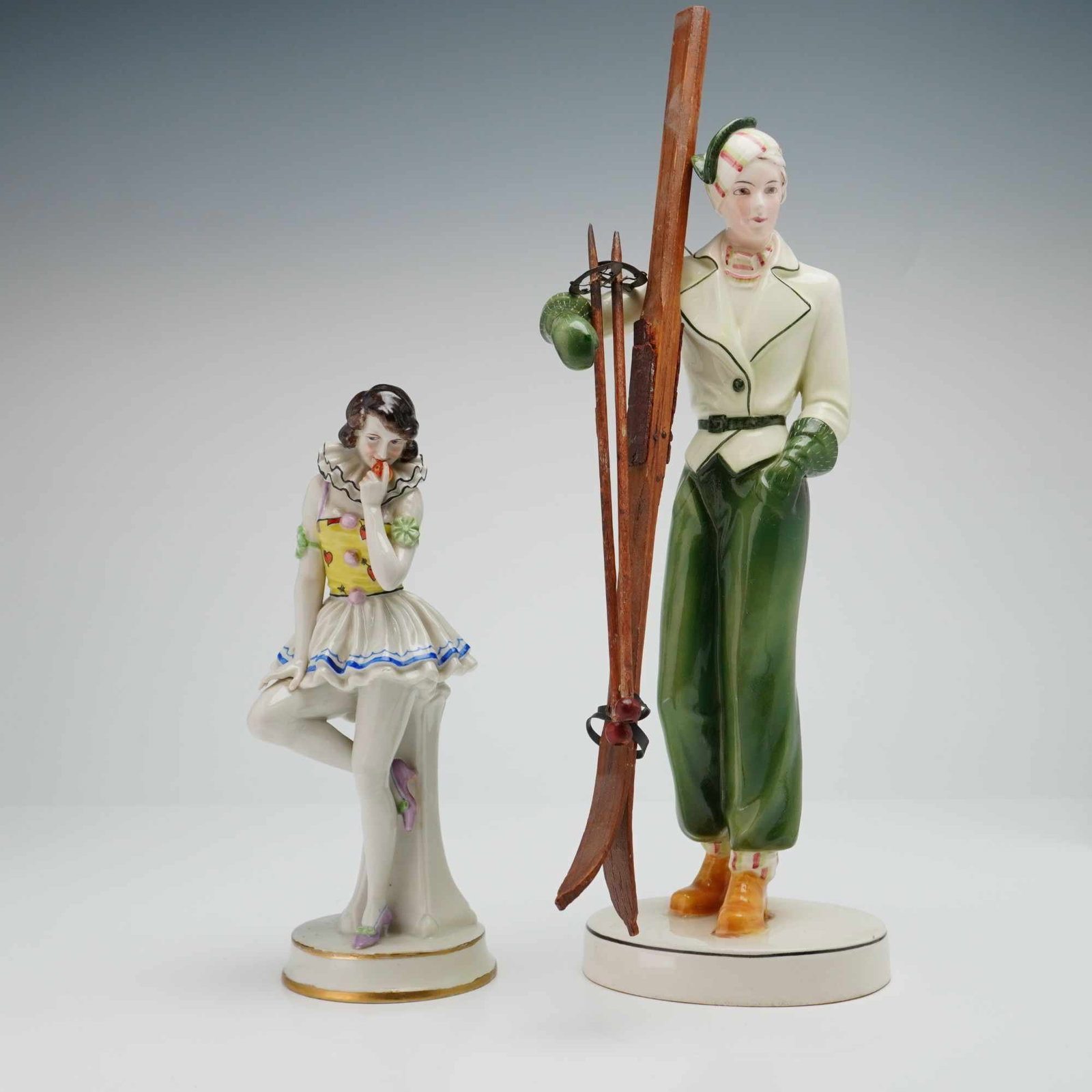2pc German Art Deco Porcelain Figurines Hertwig and Co. Skier & Volkstedt Pierette: This lot features two porcelain figurines that includes a figurine of a Pierrette and a taller sculpture of a female skier with wooden skies. The skier is identified as a work by Hertwig and Co. of Ka