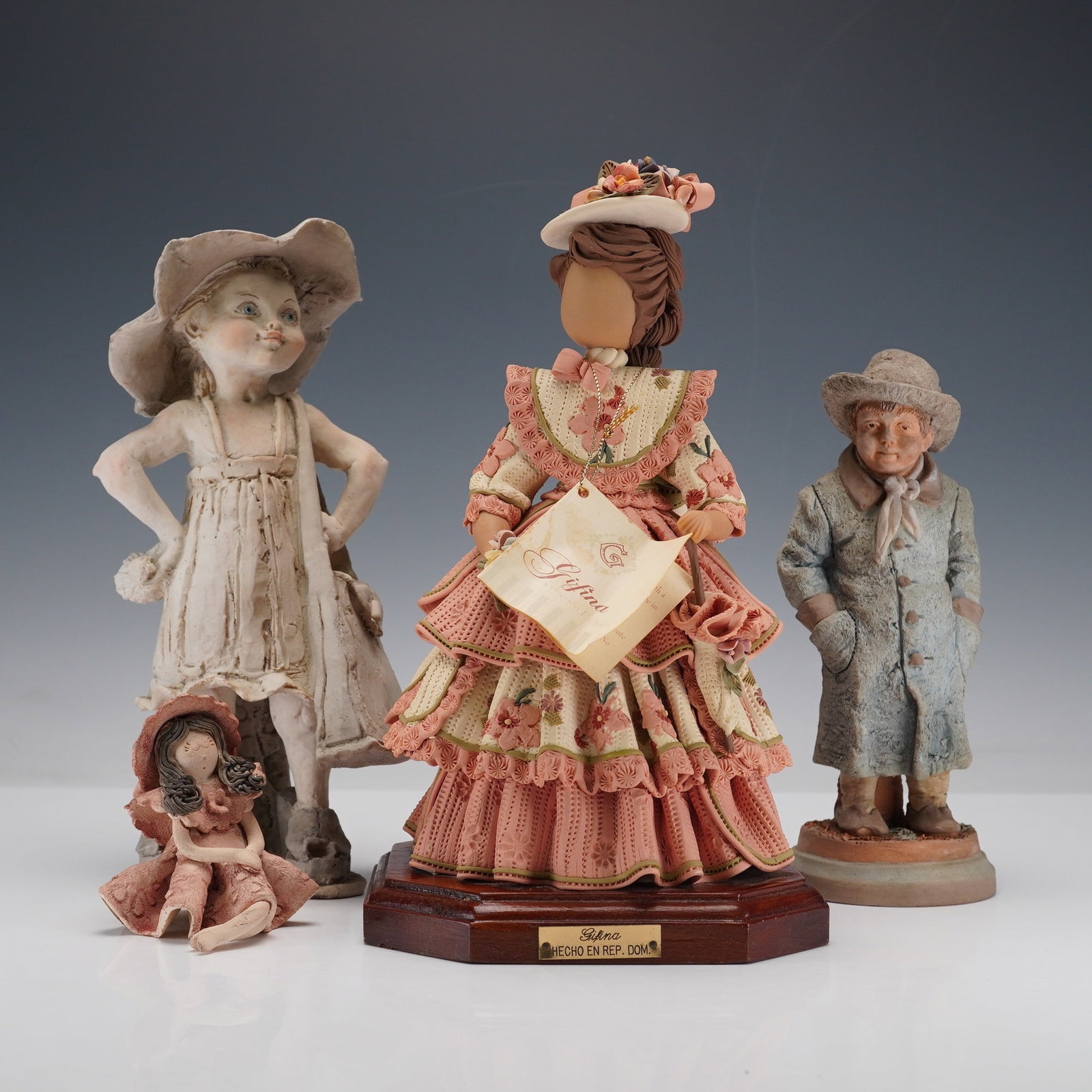 4pc Assorted Vintage Ceramic Figurines: Group of four figurines including a handcrafted ceramic figure of a lady in a layered pink lace dress with a bonnet and flowers, mounted on a wooden octagonal base with brass nameplate. Accompanying p
