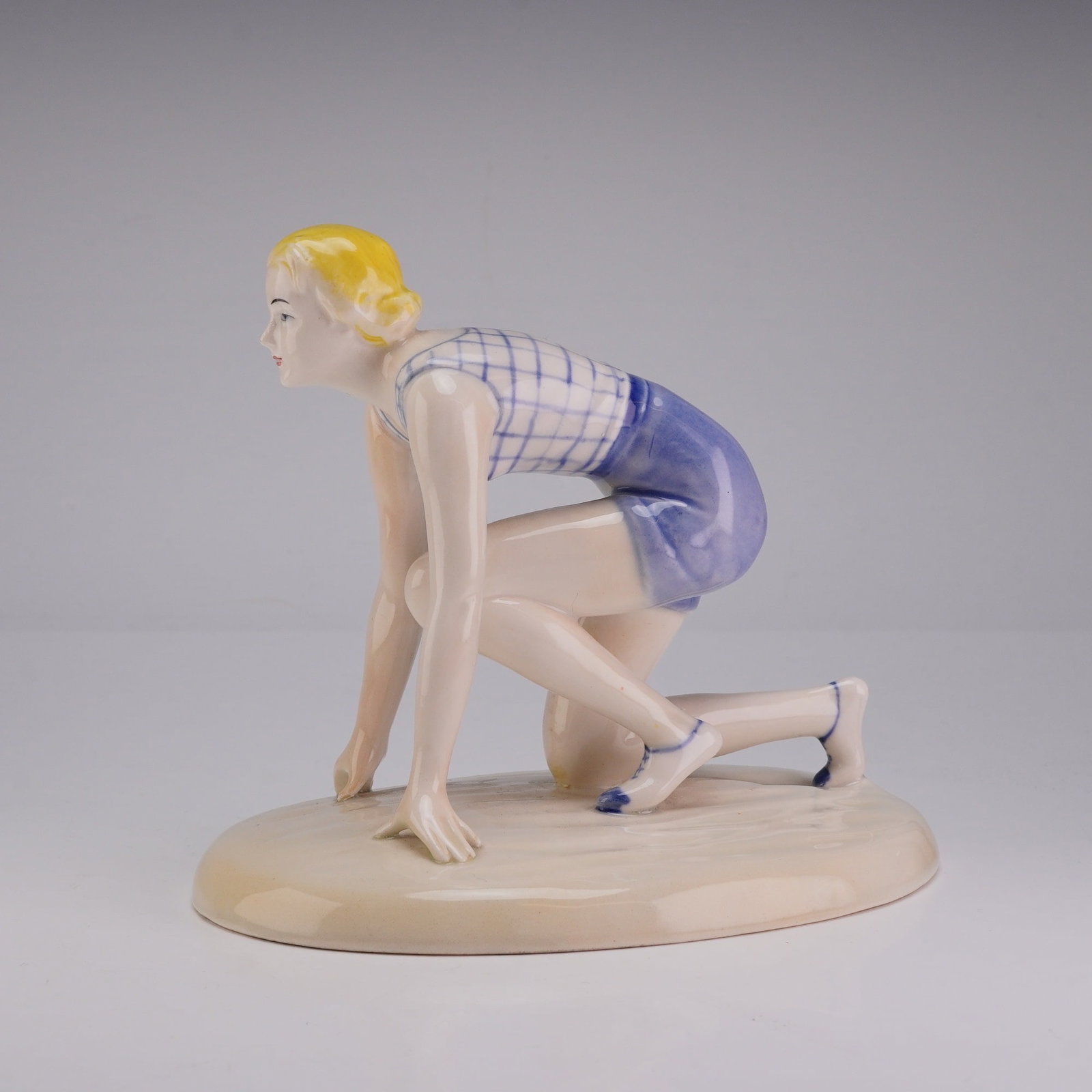 Goebel Glazed Porcelain Figurine Female Sprinter Model FF49: A Goebel glazed porcelain figurine depicting a female athlete in a starting crouch position on an integral oval base. The figure is rendered with blonde hair and wears a hand-painted blue and white ch