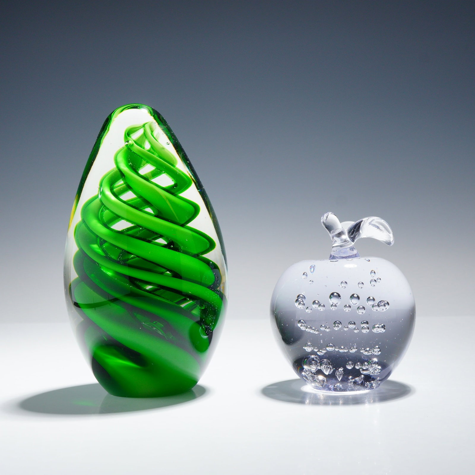 2pc Art Glass Paperweights: This lot features two contemporary art glass paperweights. The first is a tall, ovoid sculpture featuring a vibrant emerald green spiral twist core encased within a thick layer of clear glass. The bas