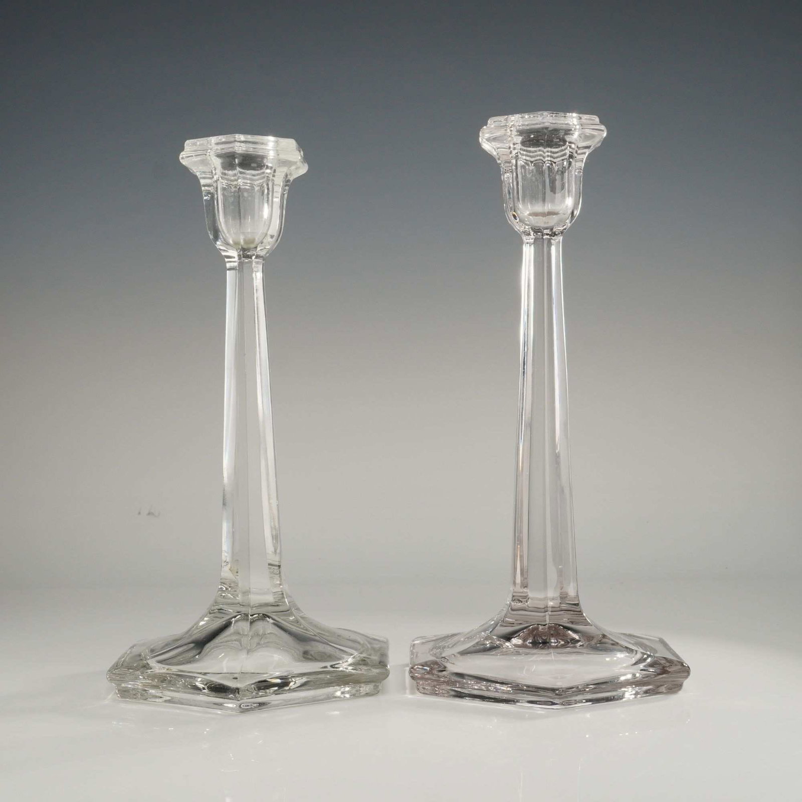 A Pair of Fostoria Clear Glass Hexagonal Candlestick Holders: A pair of crystal candlestick holders, likely Fostoria, featuring a hexagonal design. Each piece is composed of a hexagonal base and a matching tapered hexagonal stem. The candlesticks are topped with