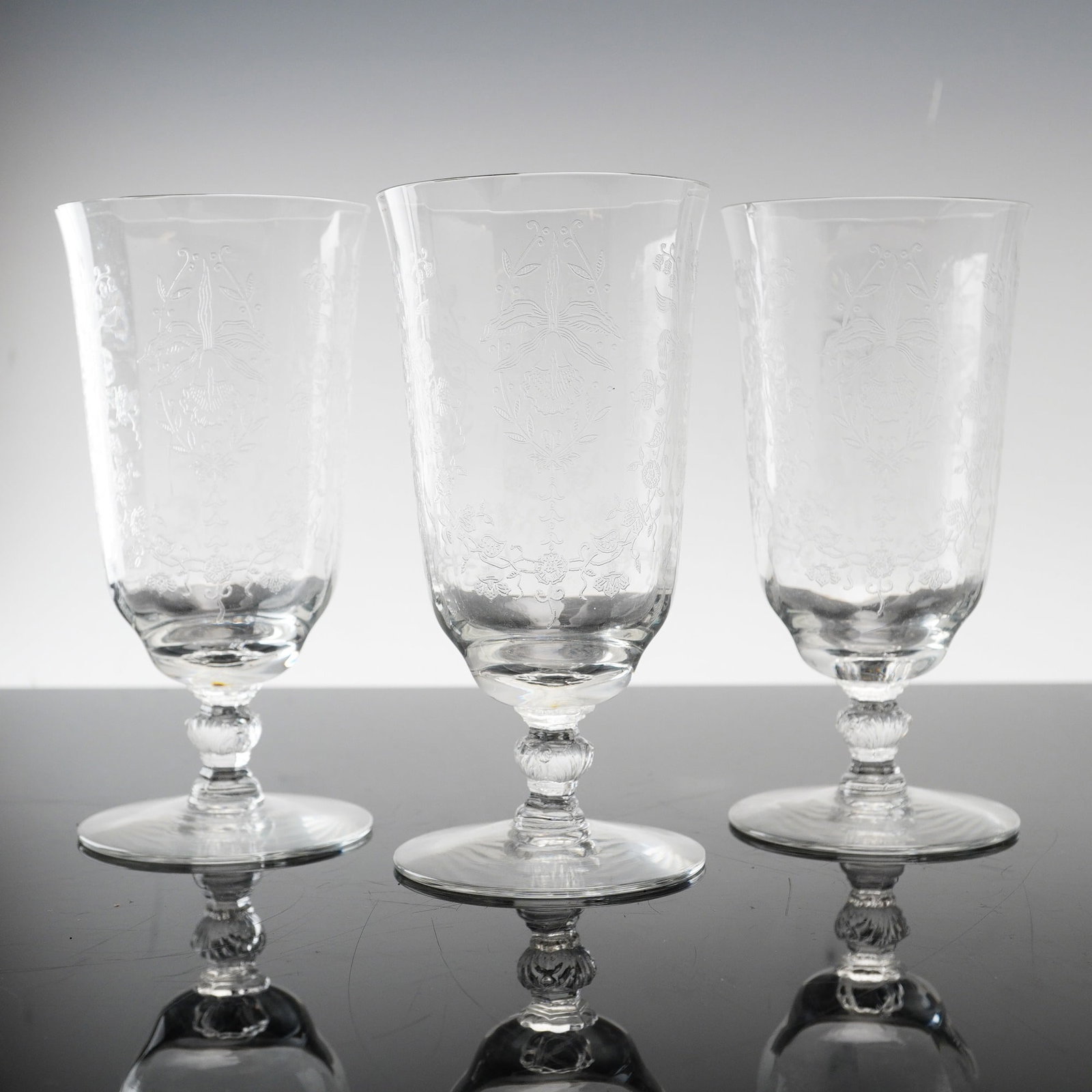 Three Heisey Etched Crystal Footed Iced Tea Glasses, Orchid Pattern: A trio of crystal footed iced tea glasses manufactured by Heisey in the Orchid pattern, etch number 507. The glasses are constructed on the 5025 stem, which features a faceted hexagonal knop and a fla