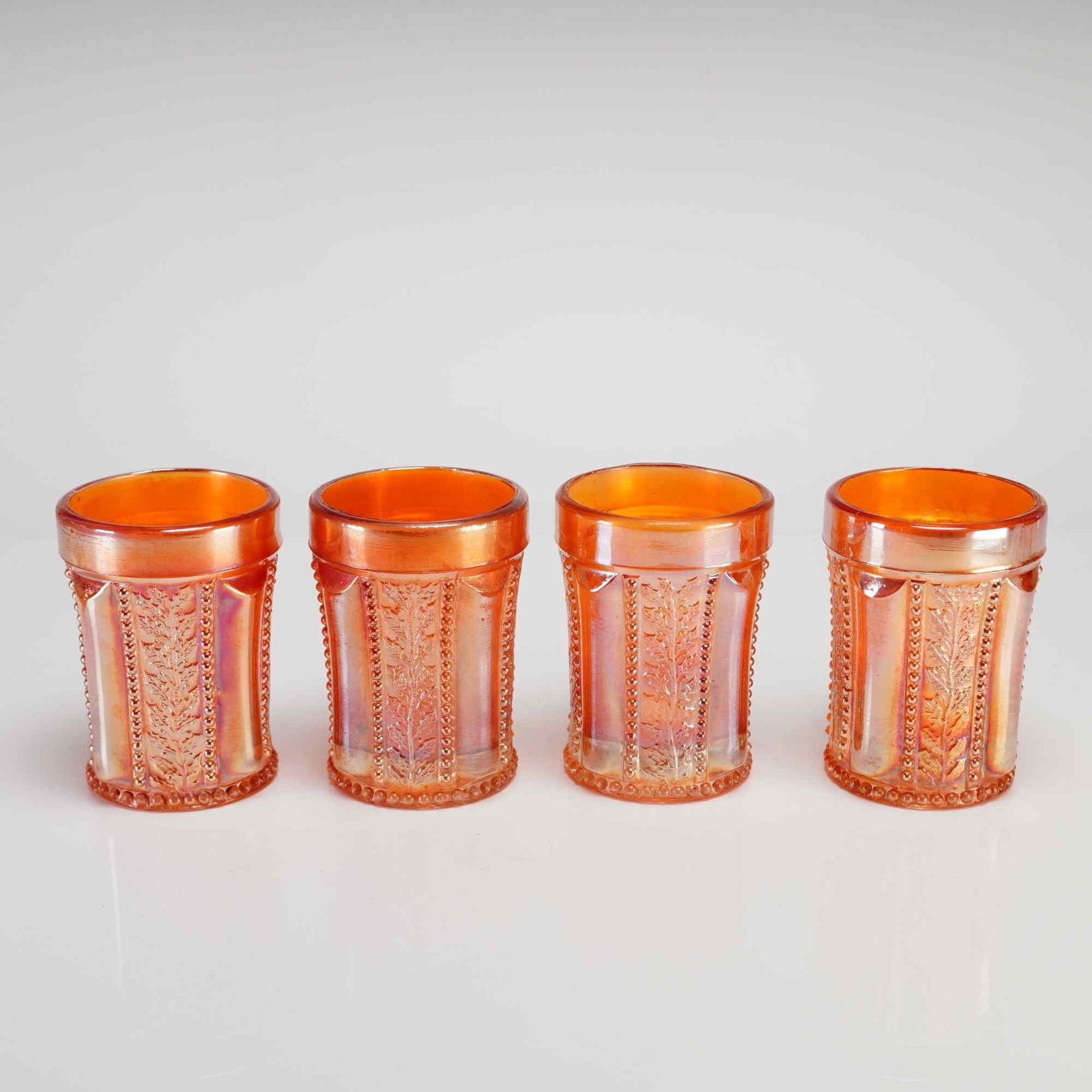 A Set of Four Marigold Carnival Glass Tumblers in the Paneled Holly Pattern: This set of four matching juice glasses is made of marigold carnival glass featuring the Paneled Holly pattern. The design consists of alternating vertical panels where one panel is smooth and termina