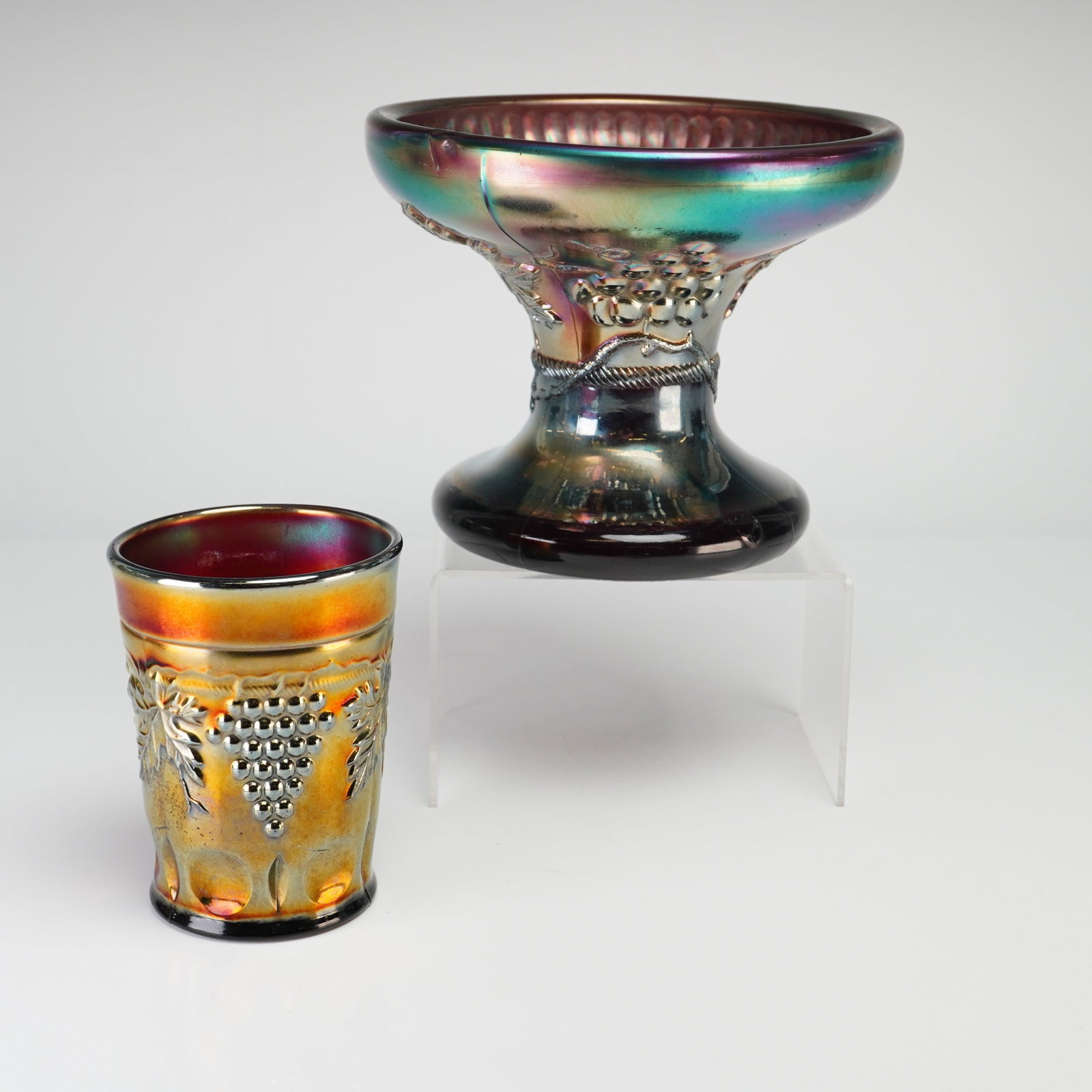Two Northwood Grape and Cable Pattern Carnival Glass Items: This lot features two pieces of American carnival glass in the Grape and Cable pattern. The first item is a pedestal compote with an iridescent finish over an amethyst base glass, displaying a spectru