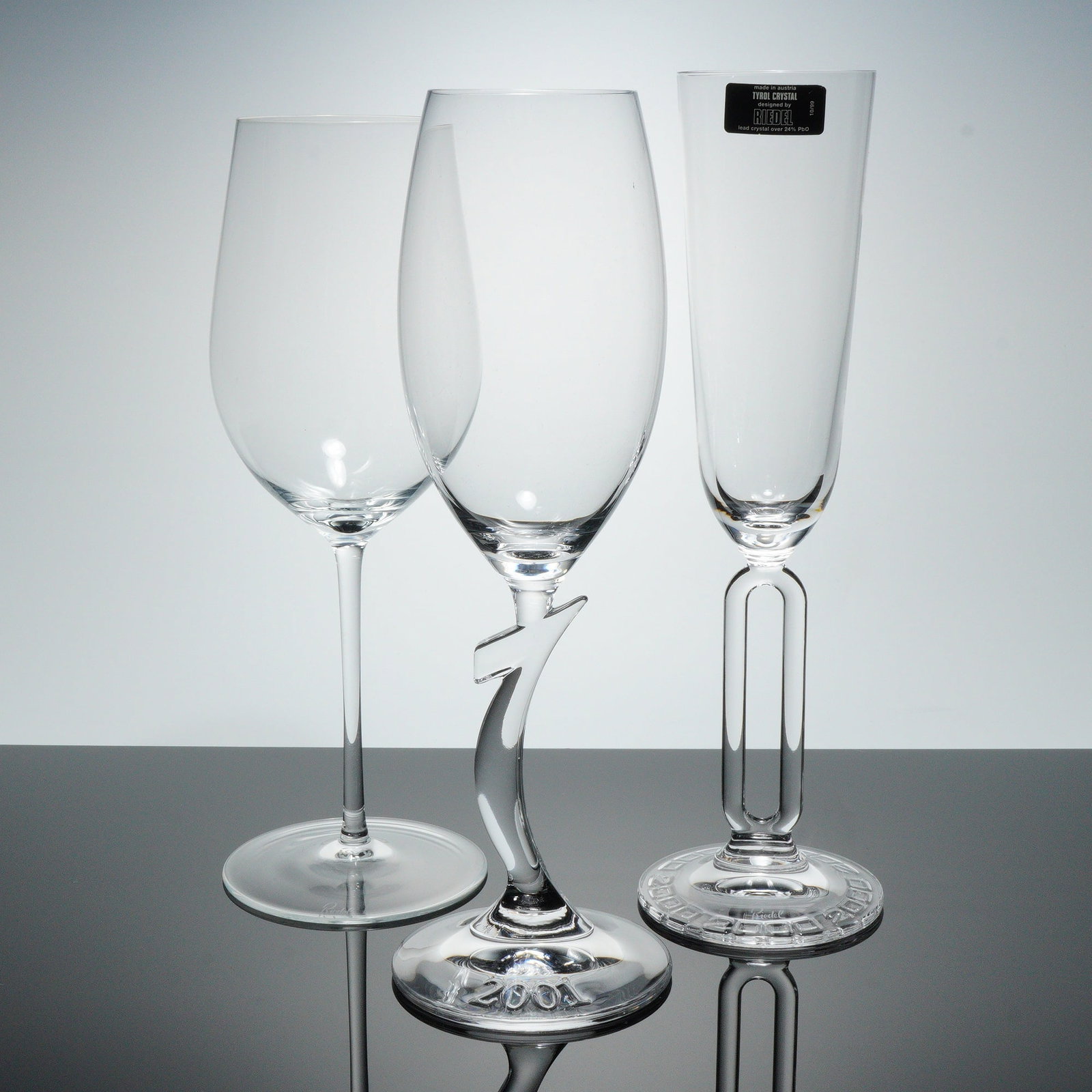 3pc Riedel Crystal Wine and Champagne Glasses: This assortment includes three pieces of clear Riedel crystal stemware. One fluted champagne glass has a hollow column stem, a foot molded with the date 2000 around the edge, and label. A second stem 