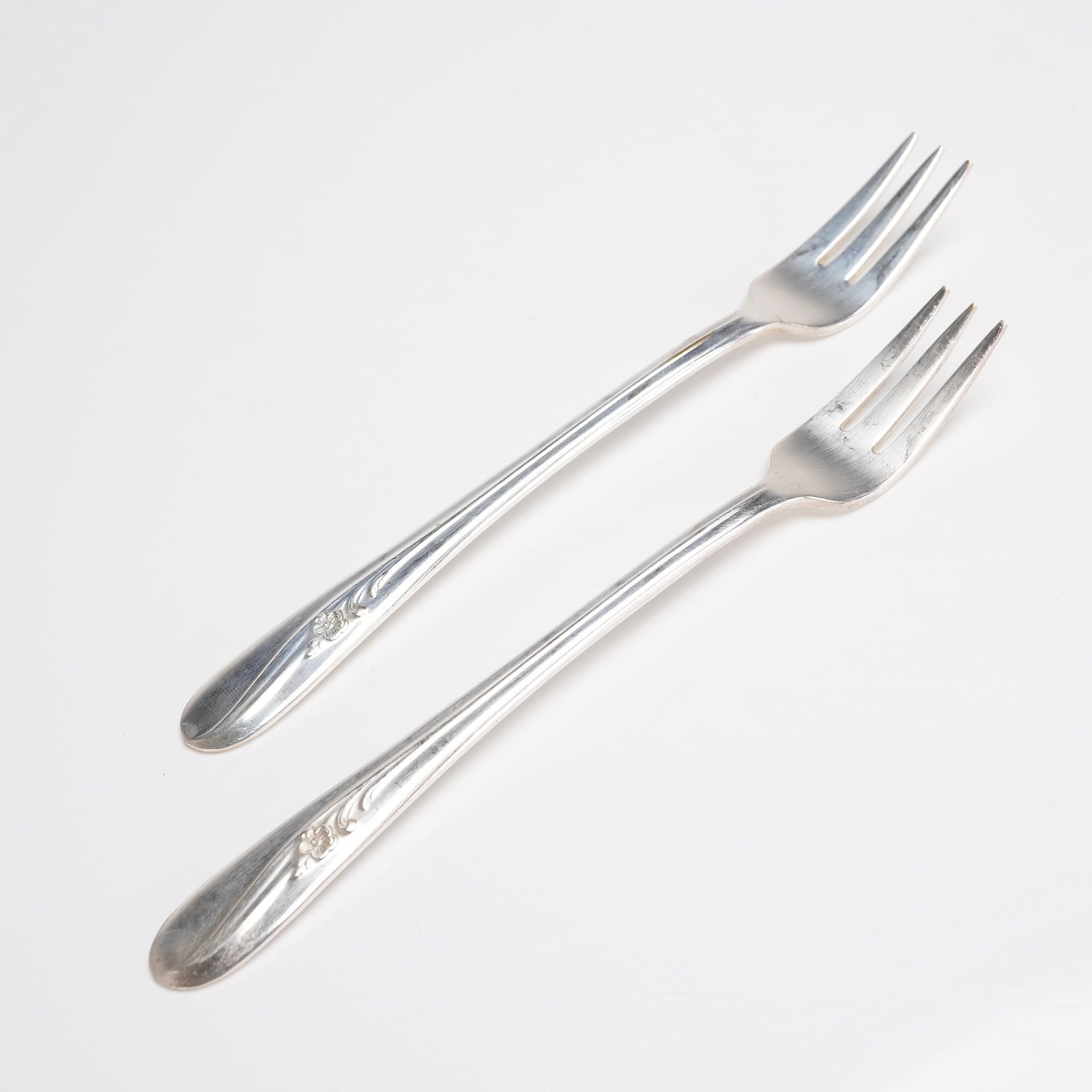 A Pair of Holmes & Tuttle Meadow Flower Silverplate Cocktail Forks: A pair of silver-plated cocktail or dessert forks manufactured by Holmes & Tuttle Manufacturing Company (H. & T. MFG. CO.) in the Meadow Flower pattern. Each piece is constructed of silver-pla