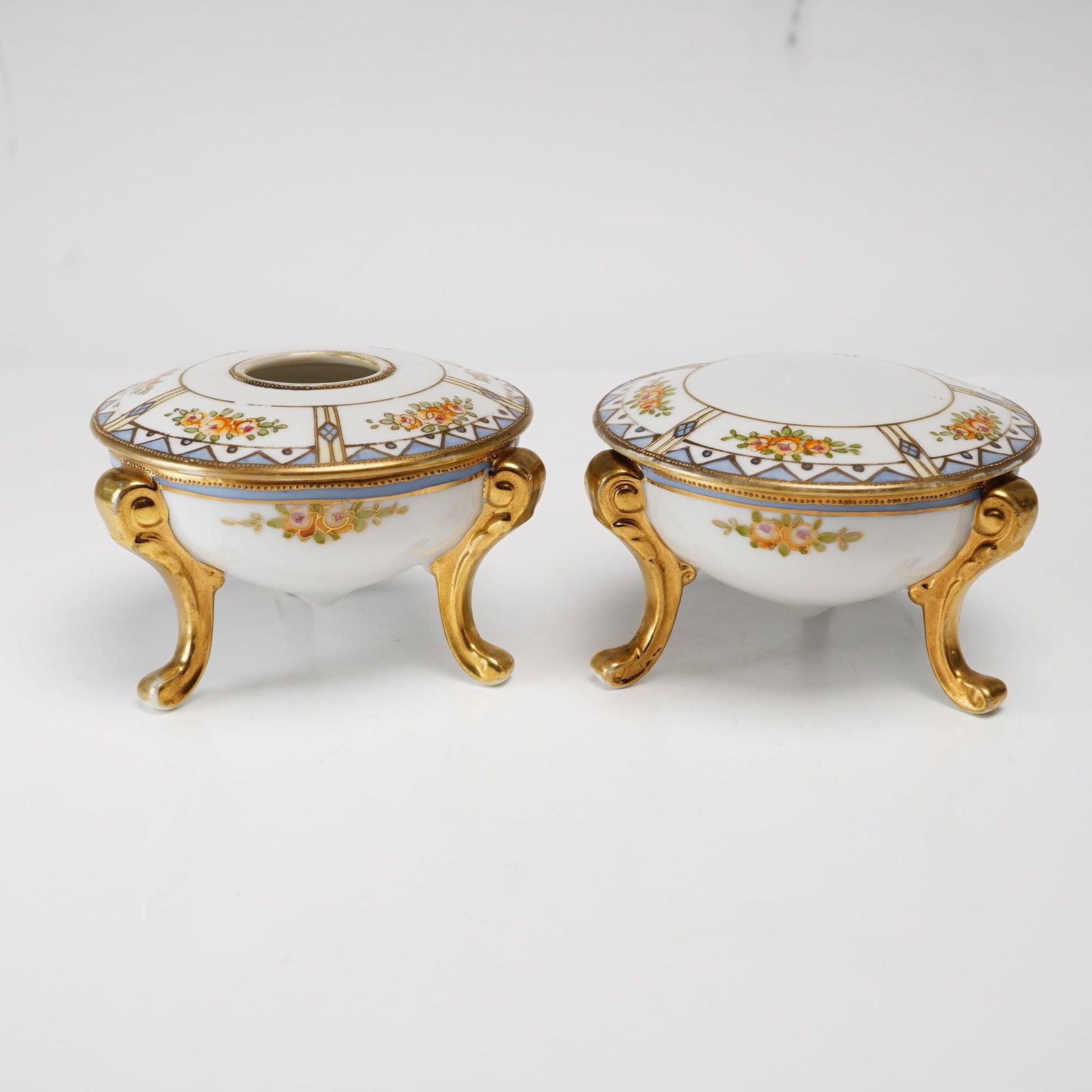 Pair of Hand Painted Nippon Porcelain Three-Footed Dresser Jars: This pair of hand-painted porcelain dresser jars consists of a powder jar and a matching hair receiver, produced by Morimura Brothers for the Nippon export market. Each piece features a bulbous, tripa
