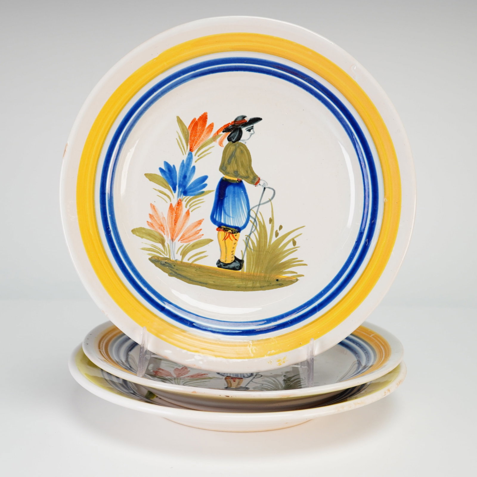 Trio of Henriot Quimper French Earthenware Faience Breton Man Plates: This trio of French faience plates is produced by the Henriot Quimper factory. Each plate is constructed of glazed earthenware and features the traditional Petit Breton or Breton Man pattern. The cent