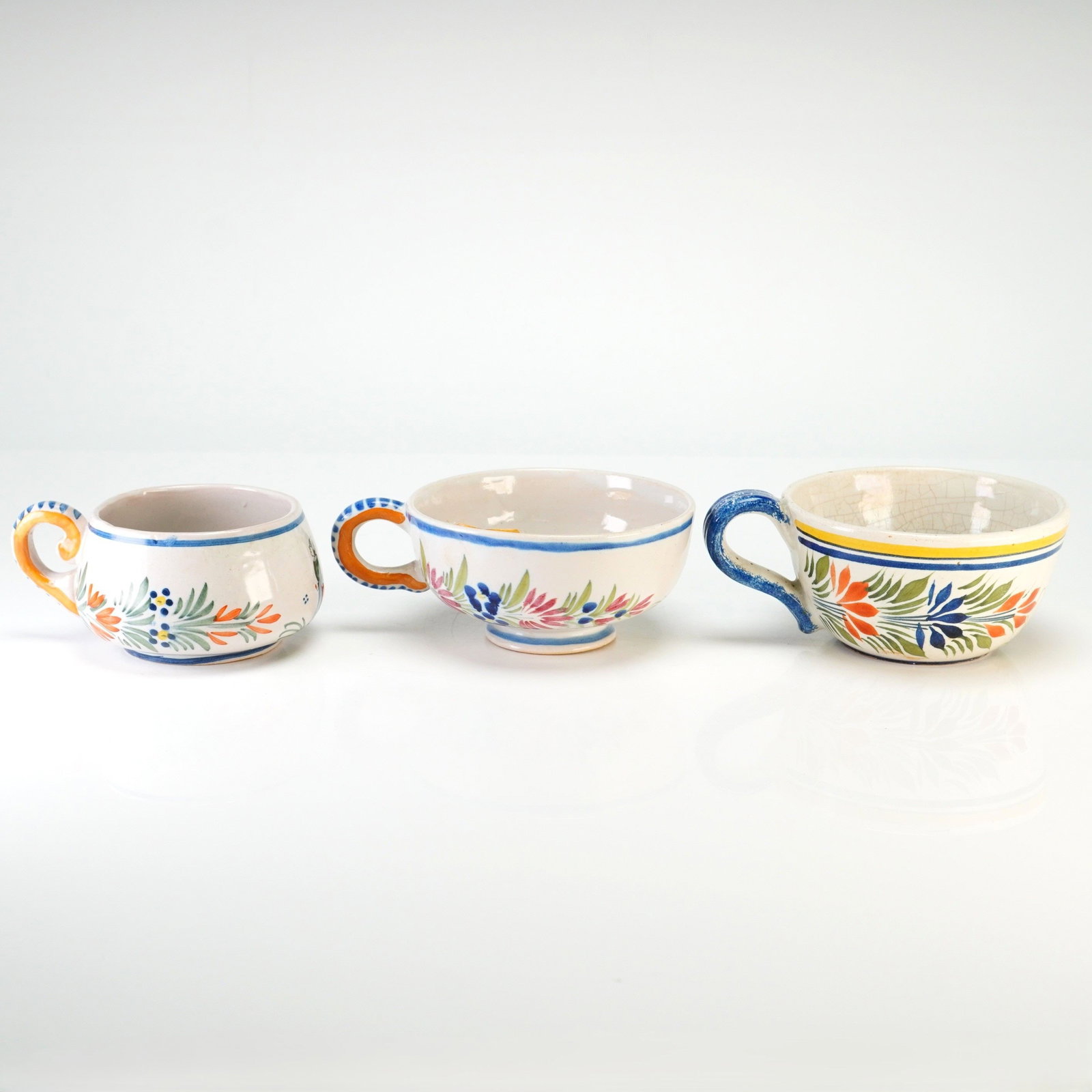 Three Piece Henriot Quimper French Earthenware Faience Cups: Each cup is constructed of glazed earthenware and features the traditional Petit Breton or Breton Man/Woman pattern. The rims are finished with characteristic concentric yellow and blue bands. The und