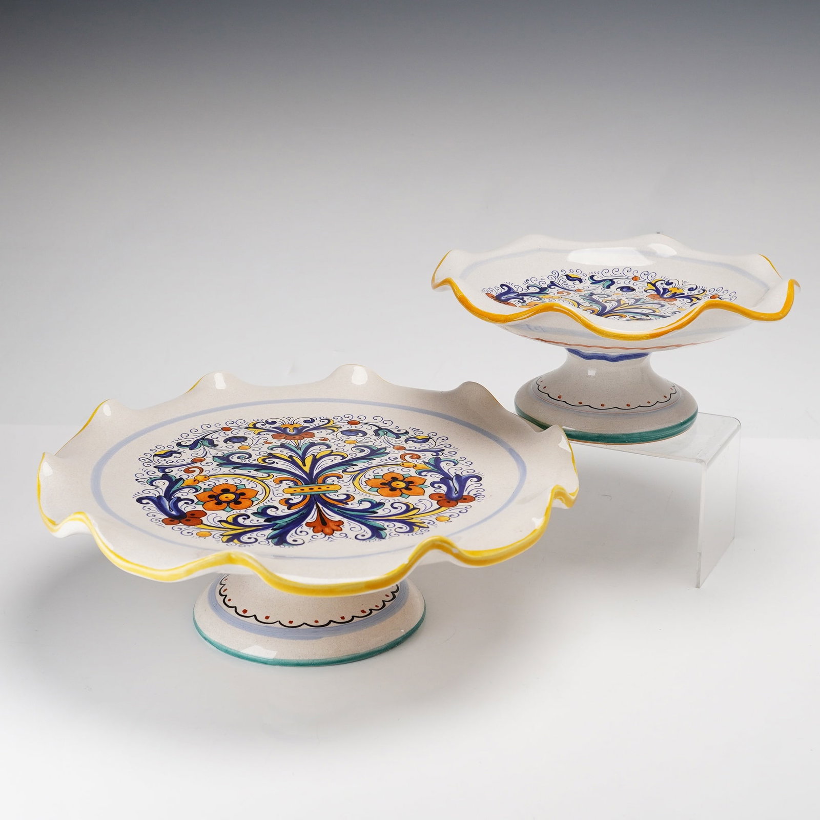 Two Piece Deruta Ceramiche Italian Ricco Style Pedestal Plates: This pair of graduated pedestal compotes is manufactured by Deruta Ceramiche in Italy. The earthenware pieces are decorated in the majolica tradition, featuring a hand-painted pattern of scrolling aca