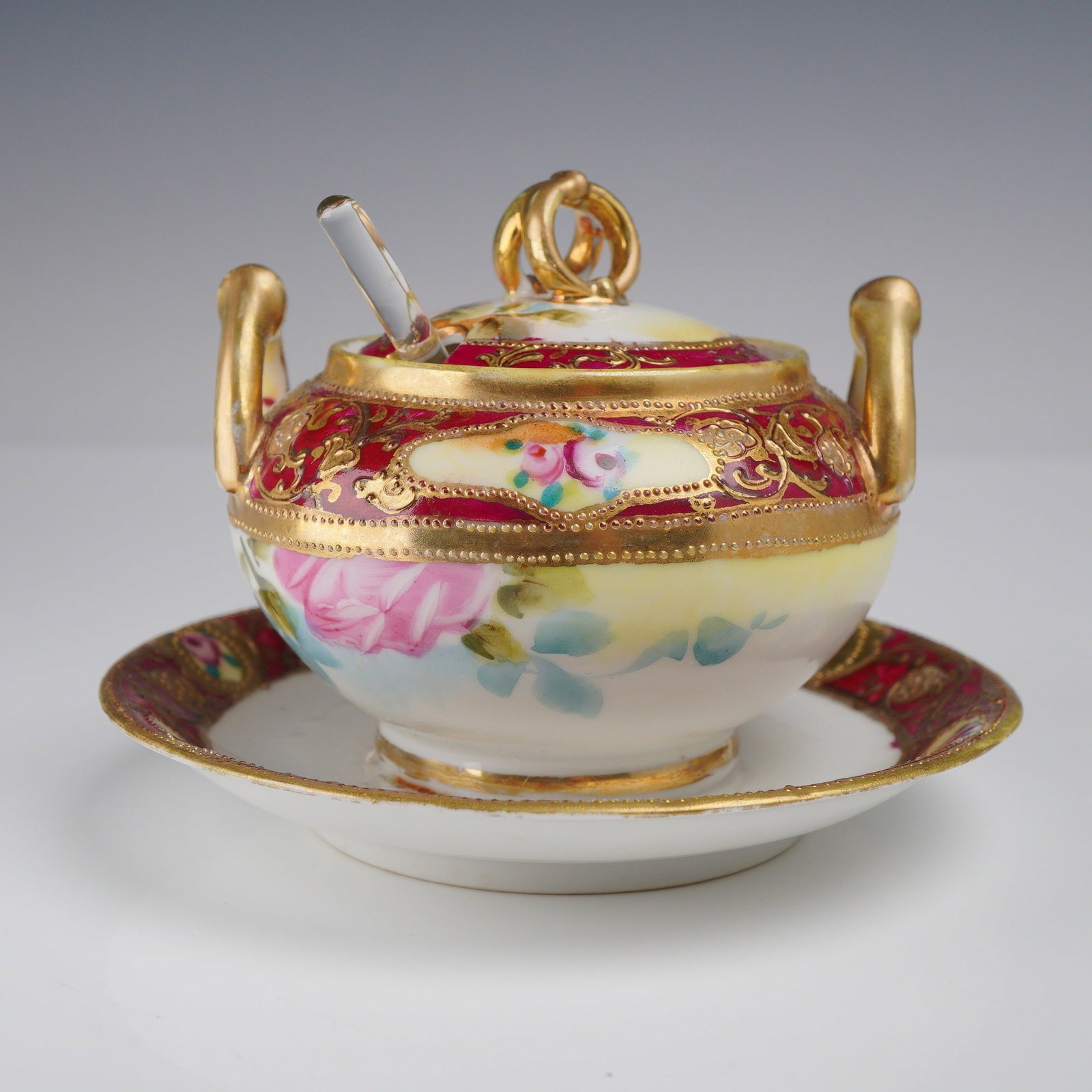 Nippon Hand-Painted Porcelain Condiment Pot with Glass Spoon: This Nippon hand-painted porcelain condiment set is a classic example of Japanese export porcelain, featuring an attached underplate, matching lid, and a clear glass spoon. The set is adorned with lus