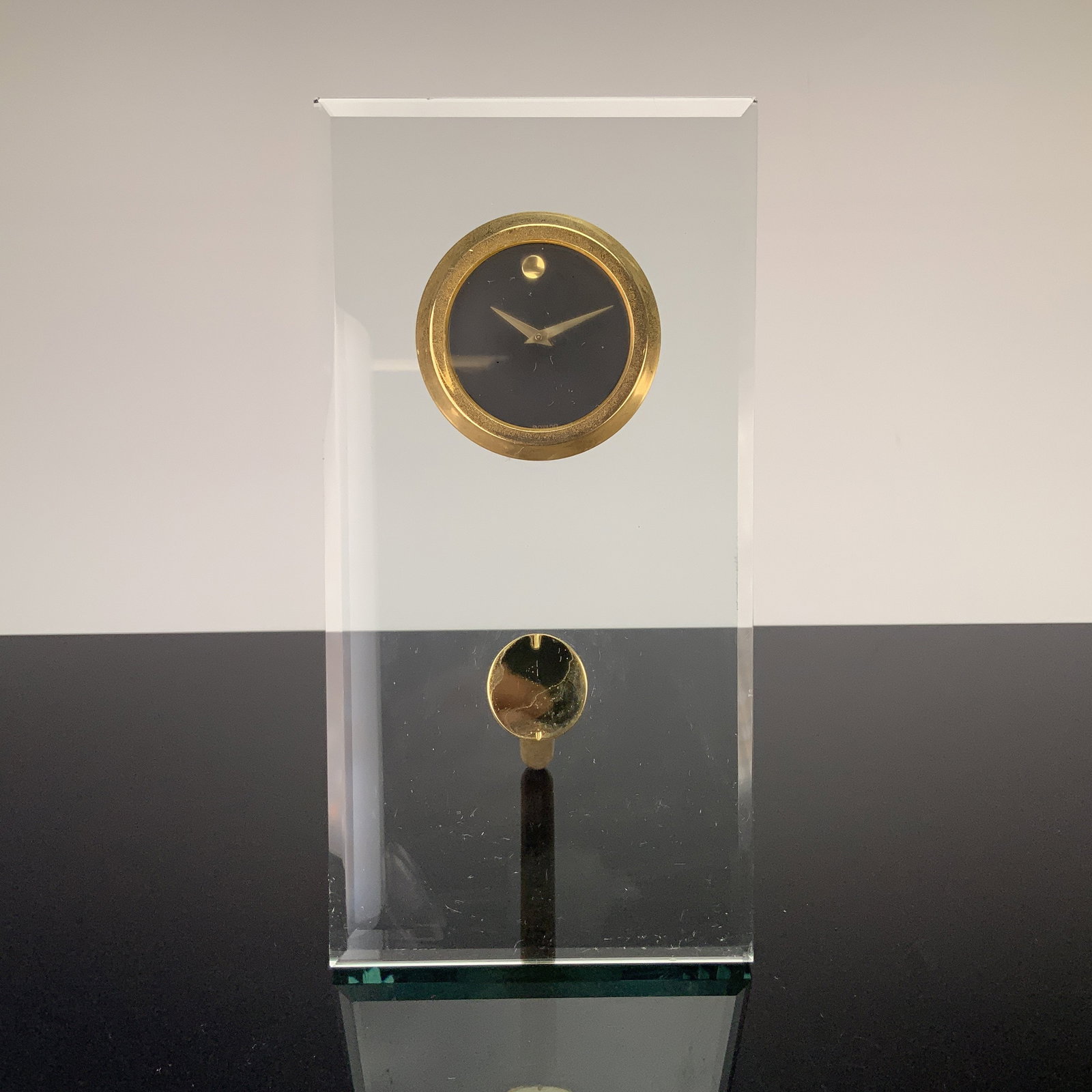 Movado Dial Cut Crystal Desk Clock: This Movado dial desk clock, features a rectangular panel of beveled clear crystal. The clock face displays a black dial, a single gold-tone dot at the 12 o'clock position, and two gold-tone tapered h