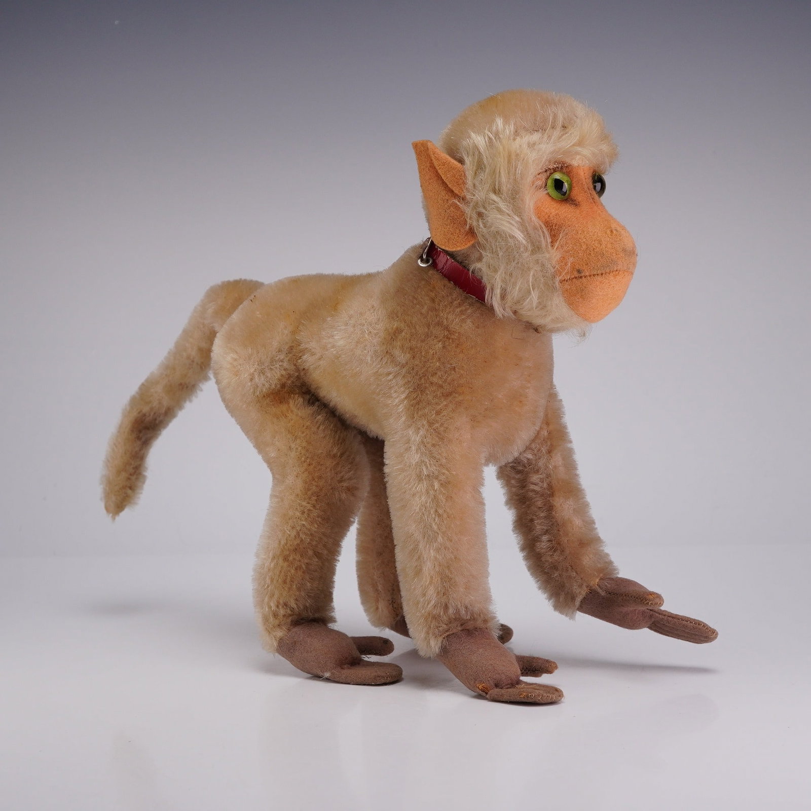 Steiff Coco Baboon Monkey: This vintage plush figure is depicted in a standing, four-legged pose. The body is constructed from light tan plush fabric, featuring a longer, shaggy blonde mane surrounding the face. The face and ea