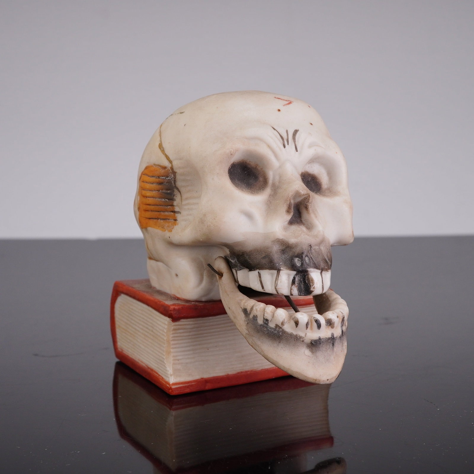 Japanese Bisque Skull on Books Nodder w/ Moving Jaw & Hole on Top: This mid-20th century Japanese bisque porcelain novelty figurine features a skull resting on books. The item is designed as a nodder with a moving jaw attached by metal pins. A circular hole on top of