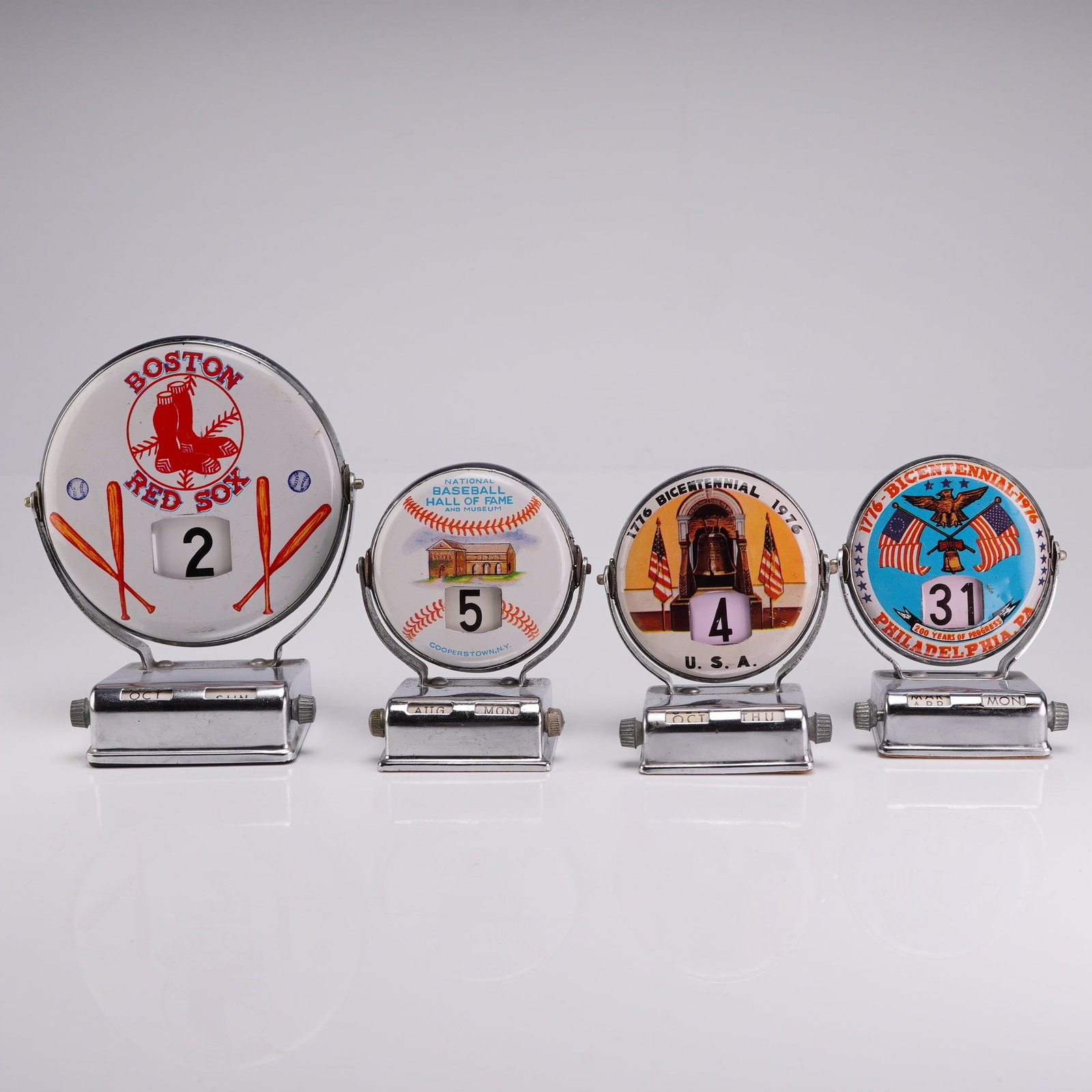 A Collection of 4 Vintage Metal Roll Top Perpetual Flip Desk Calendars: A collection of four vintage mechanical perpetual flip desk calendars featuring American sports and Bicentennial themes. These items are constructed from chrome-plated metal with printed face inserts.