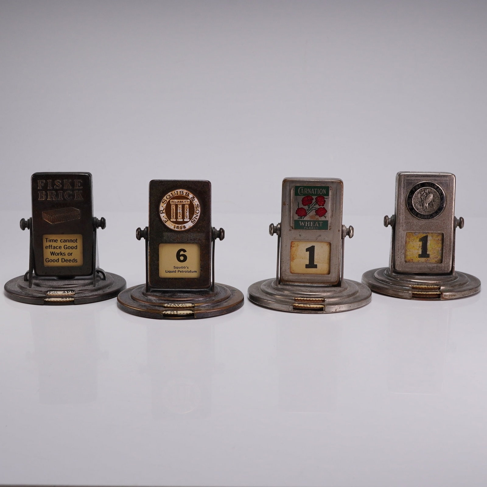 A Collection of Four Vintage Advertising Metal Roll Top Perpetual Flip Desk Calendars: This collection consists of four perpetual flip-style desk calendars, each constructed of finished metal, features a central rectangular date display that rotates on side pivots. The calendars are mou