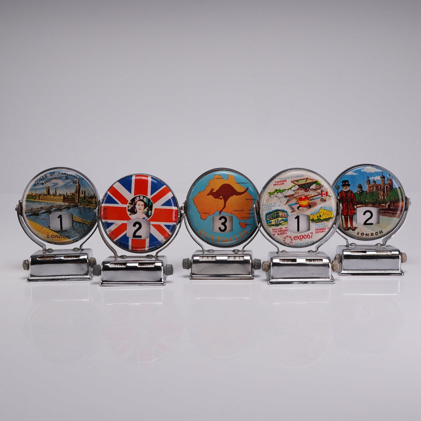 Five Vintage Souvenir Metal Flip Mechanical Roll Top Perpetual Desk Calendars: These mechanical desk accessories are constructed with chrome-plated metal bases and frames, featuring printed dials and internal scrolls. The collection includes various international souvenir themes