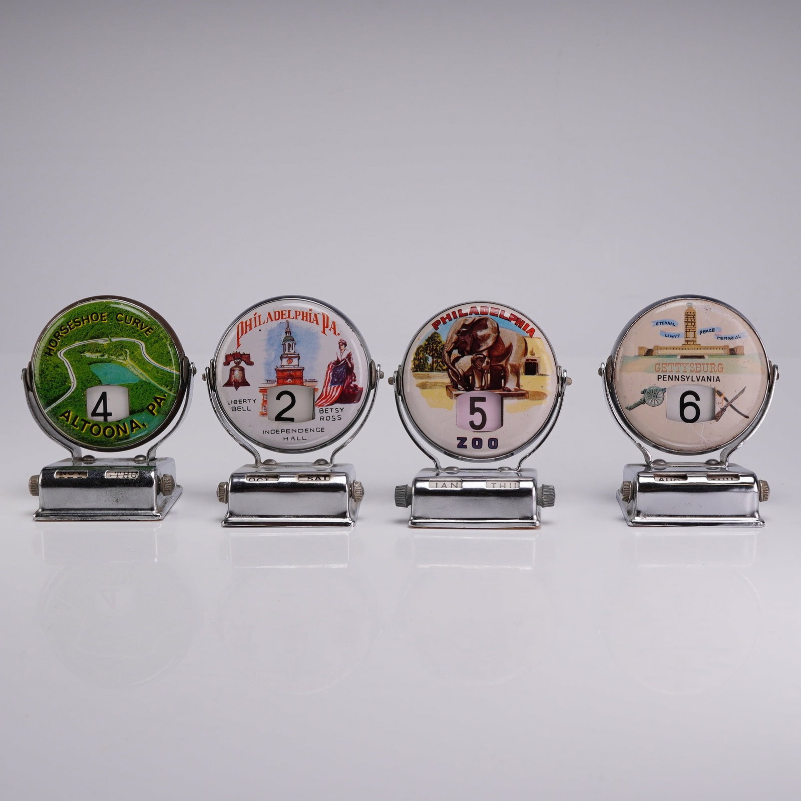 4pc Vintage Pennsylvania Souvenir Chrome Roll Top Perpetual Desk Calendars: This group of four mid-20th century perpetual desk calendars features various Pennsylvania tourist destinations. Each unit is constructed with a chrome-plated metal base and a rotating circular dial. 
