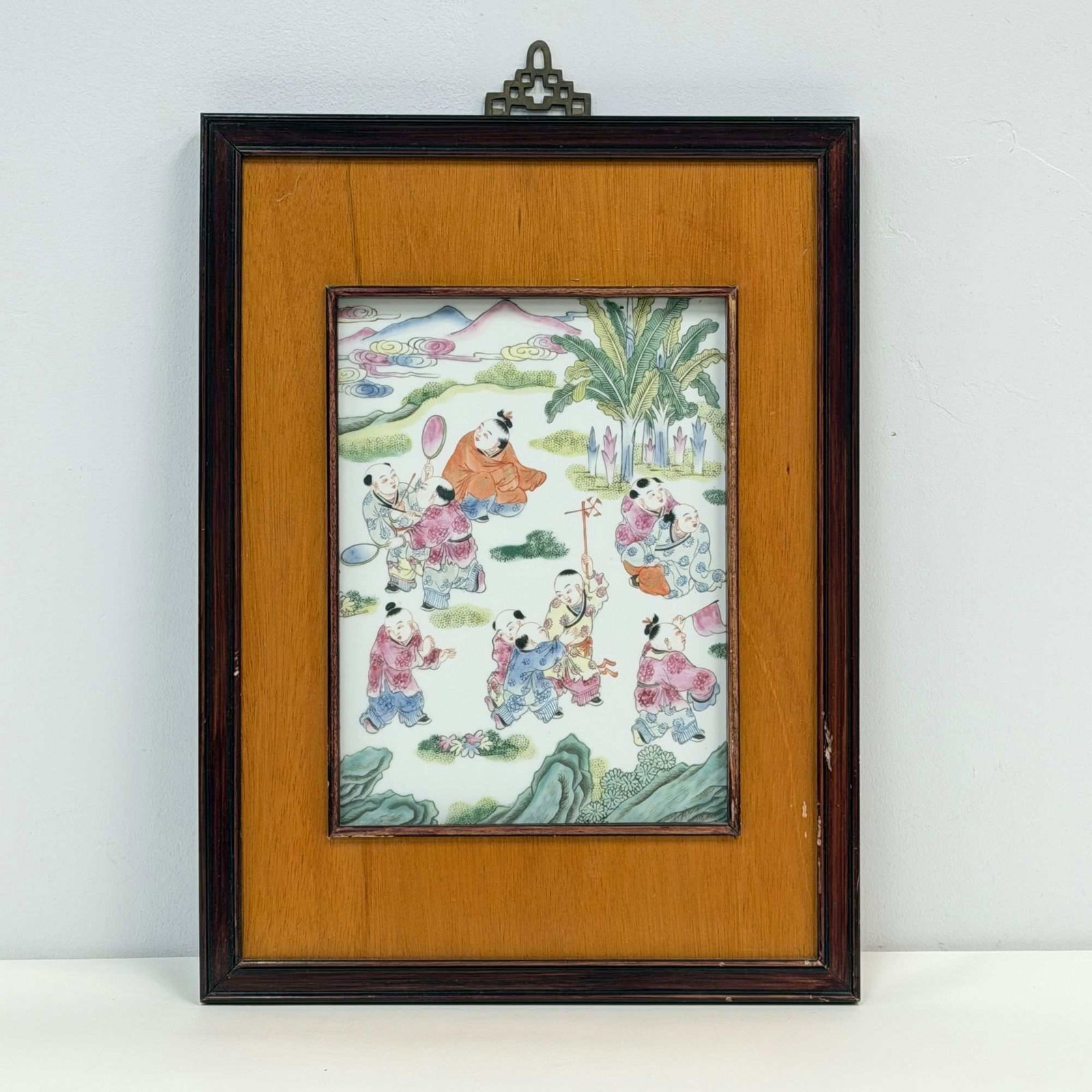 A Framed Chinese Famille Rose Porcelain Plaque Depicting Boys at Play: A rectangular Chinese porcelain plaque featuring hand painted overglaze enamel decoration in the famille rose palette. The scene depicts a traditional motif of ten children, or boys at play, engaged i