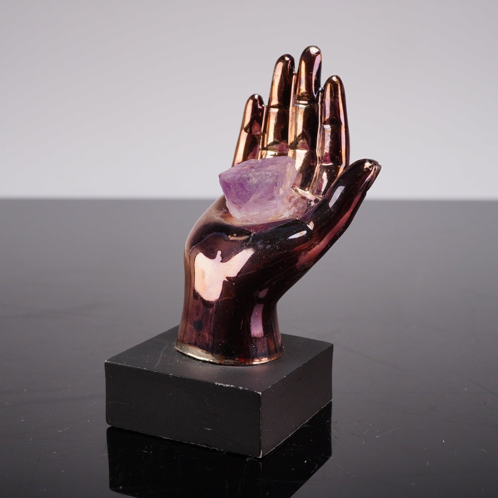 Metal Hand Sculpture on Hexagonal Base: A small figural sculpture depicting a human hand. The metal piece features a highly reflective, iridescent finish. The hand holds an amethyst-like stone. The sculpture is mounted onto a square base. S